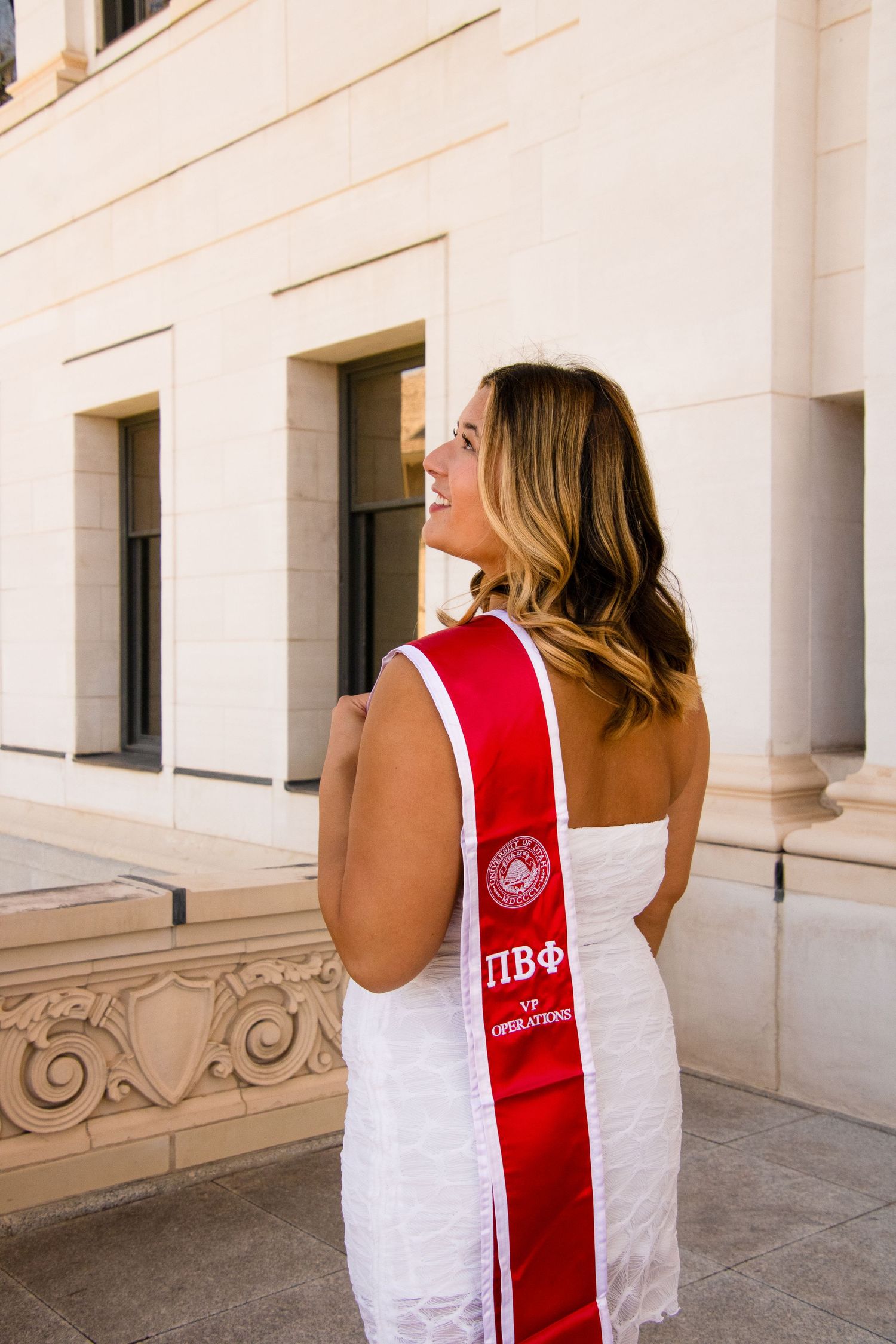 Graduate wearing white dress with red Pi Beta Phi sash poses against stone building.