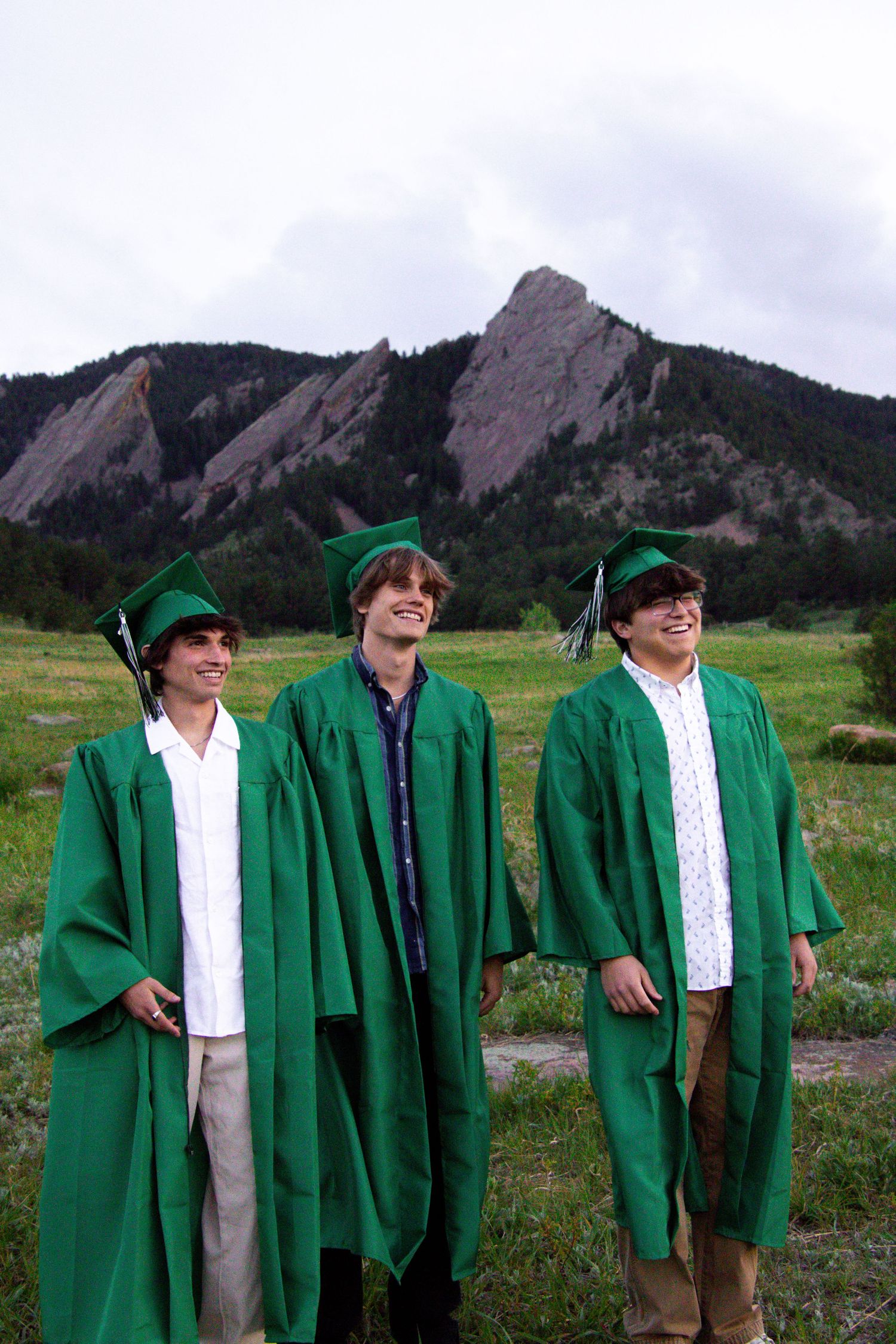 Three graduates in green graduation robes pose against mountain landscape backdrop.
