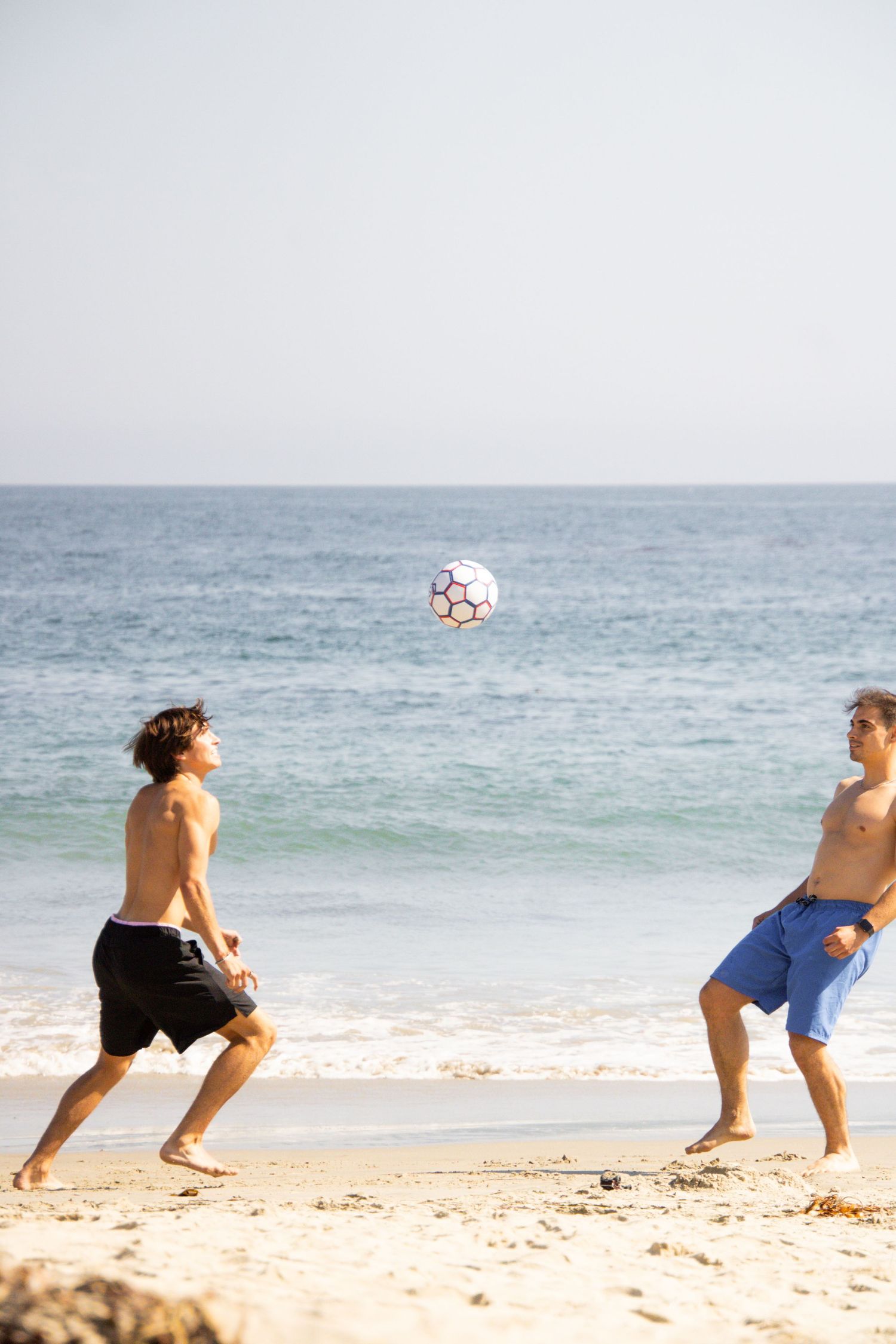 Multiple people play soccer together on a sunny beach by the ocean.