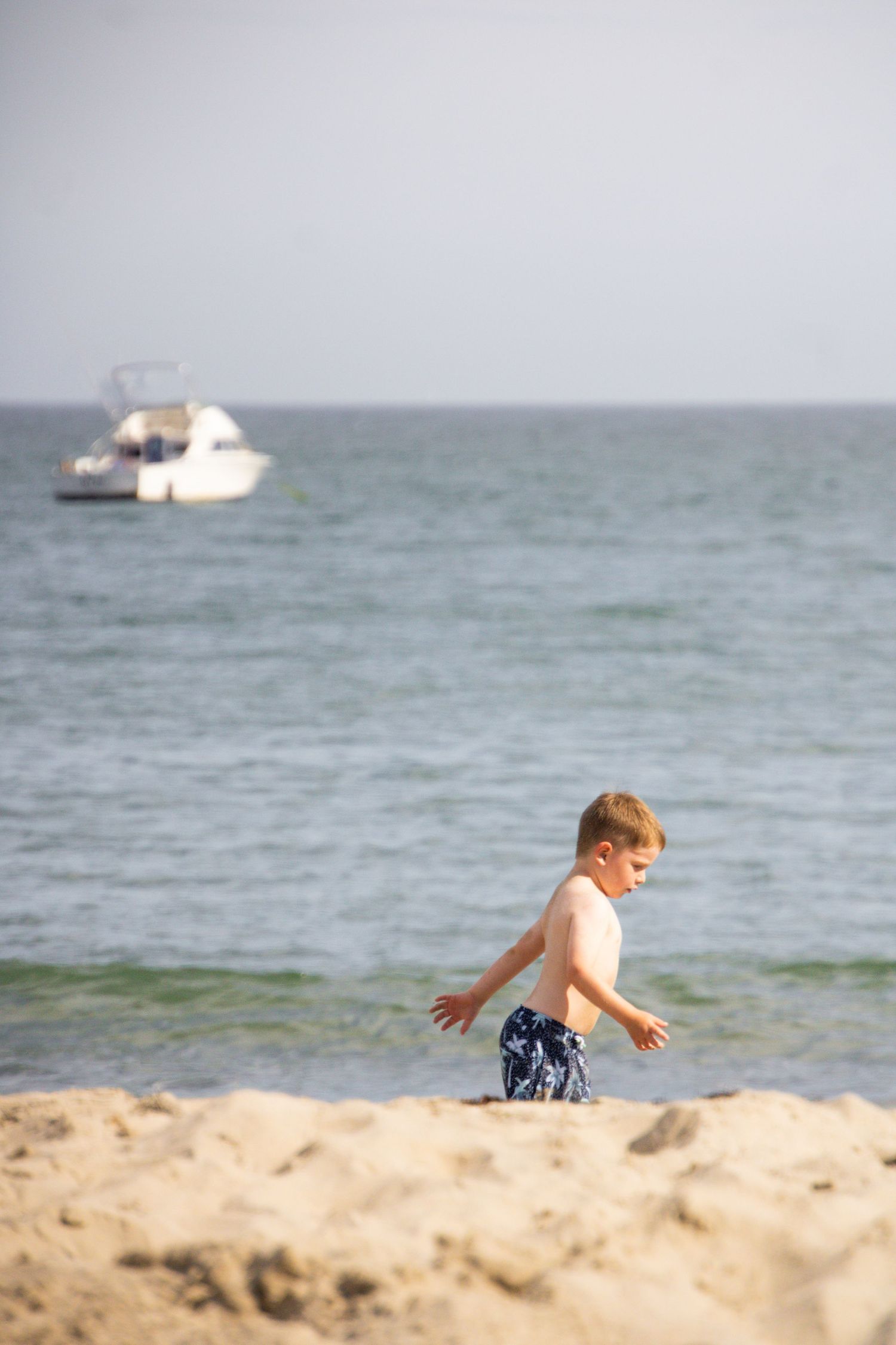 Child playing on sandy beach with boats visible in background ocean.