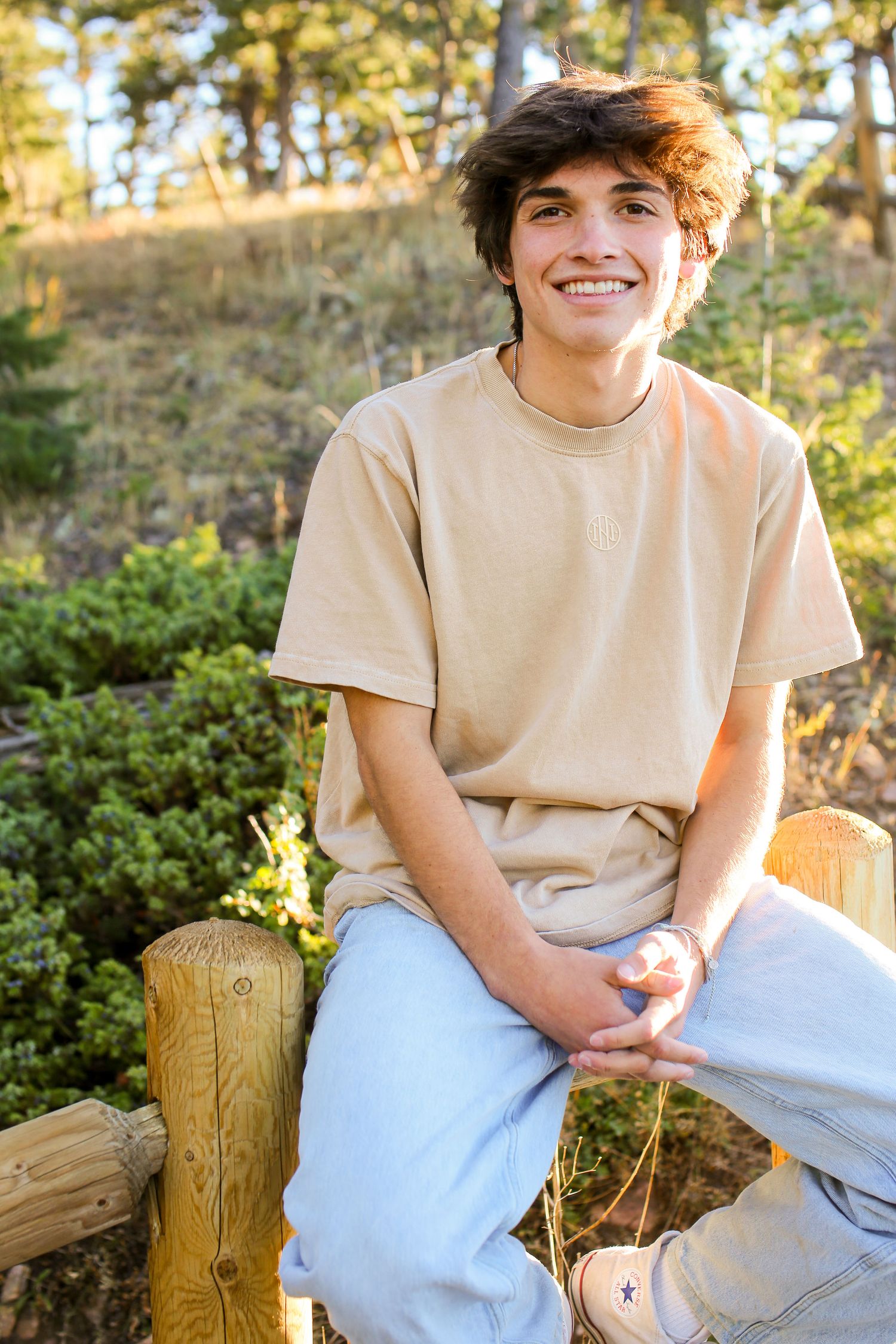 A casual outdoor portrait in warm lighting showing someone sitting on a wooden post wearing a beige t-shirt and jeans.