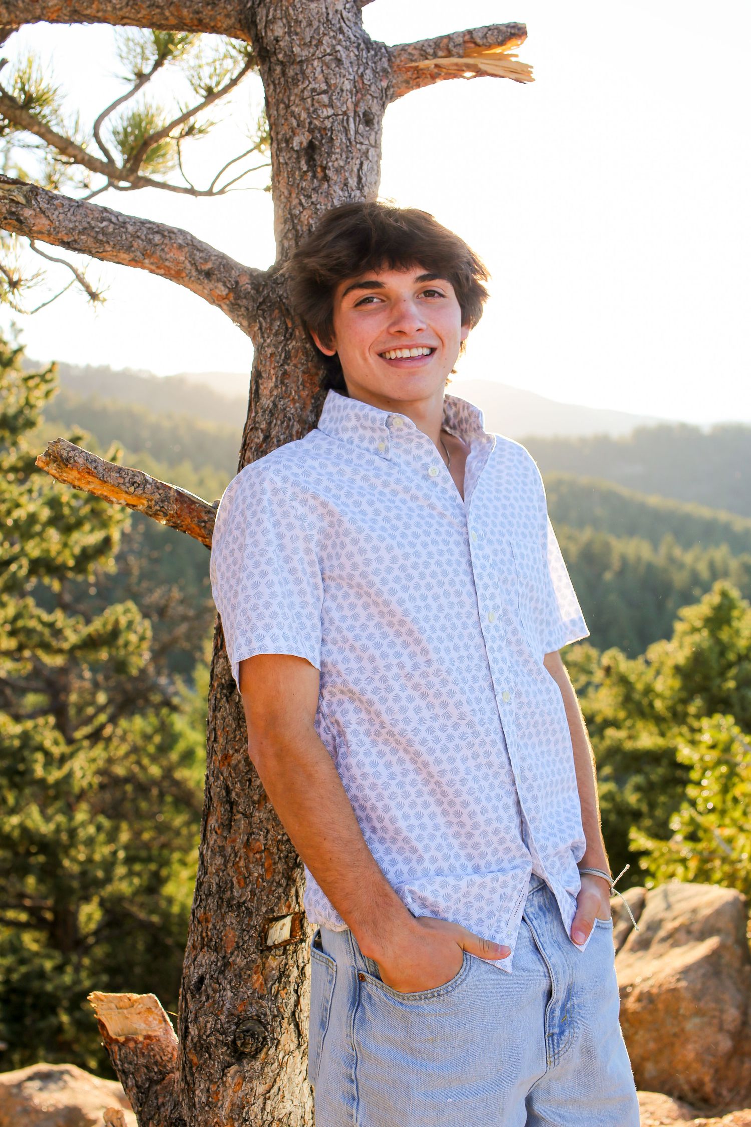 Person leaning against tree wearing polka dot shirt and jeans in golden sunlight.