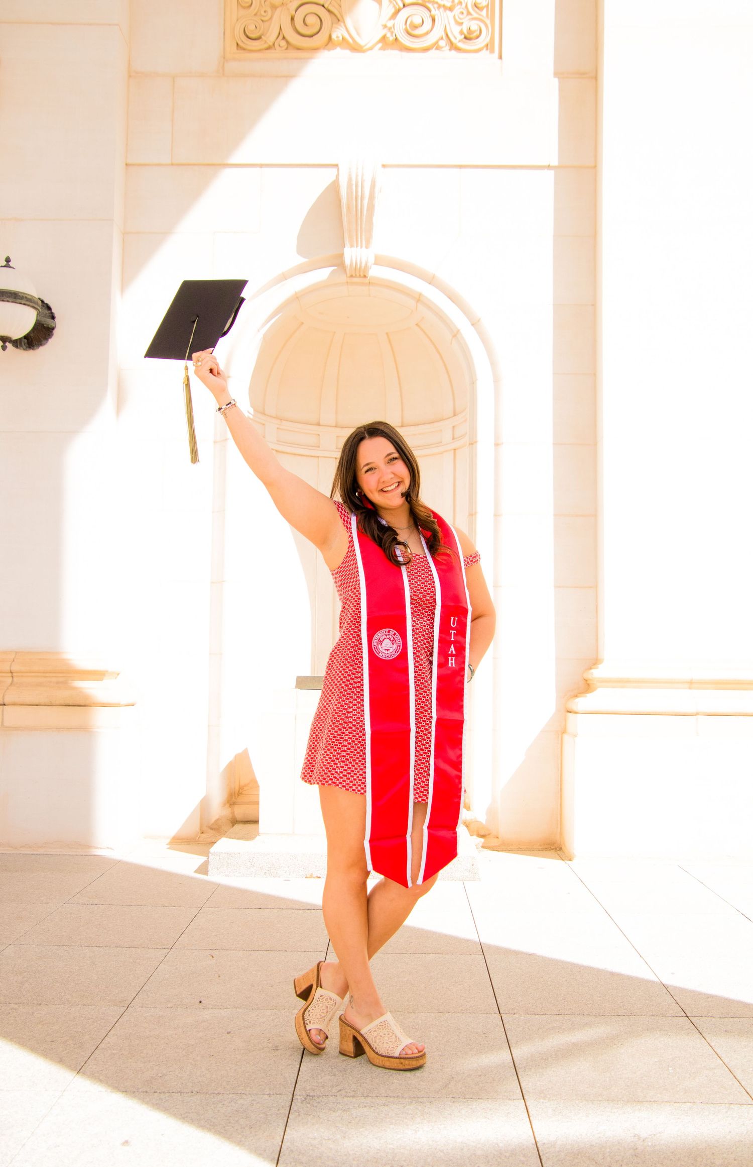 Graduate in red stole and black gown poses playfully in sunlit colonnade.