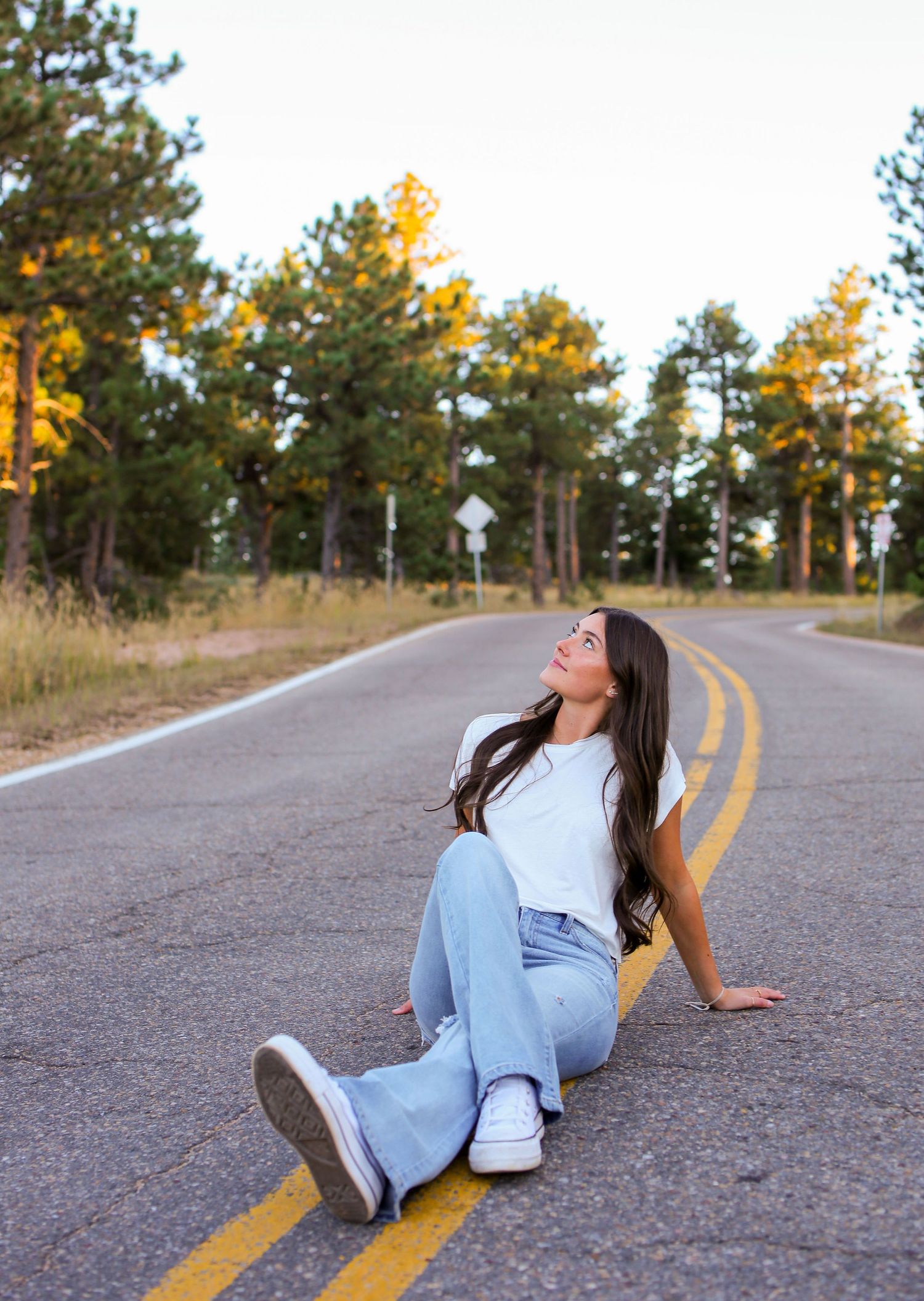 Casual portrait of someone sitting on yellow road lines wearing jeans and white sneakers at sunset.
