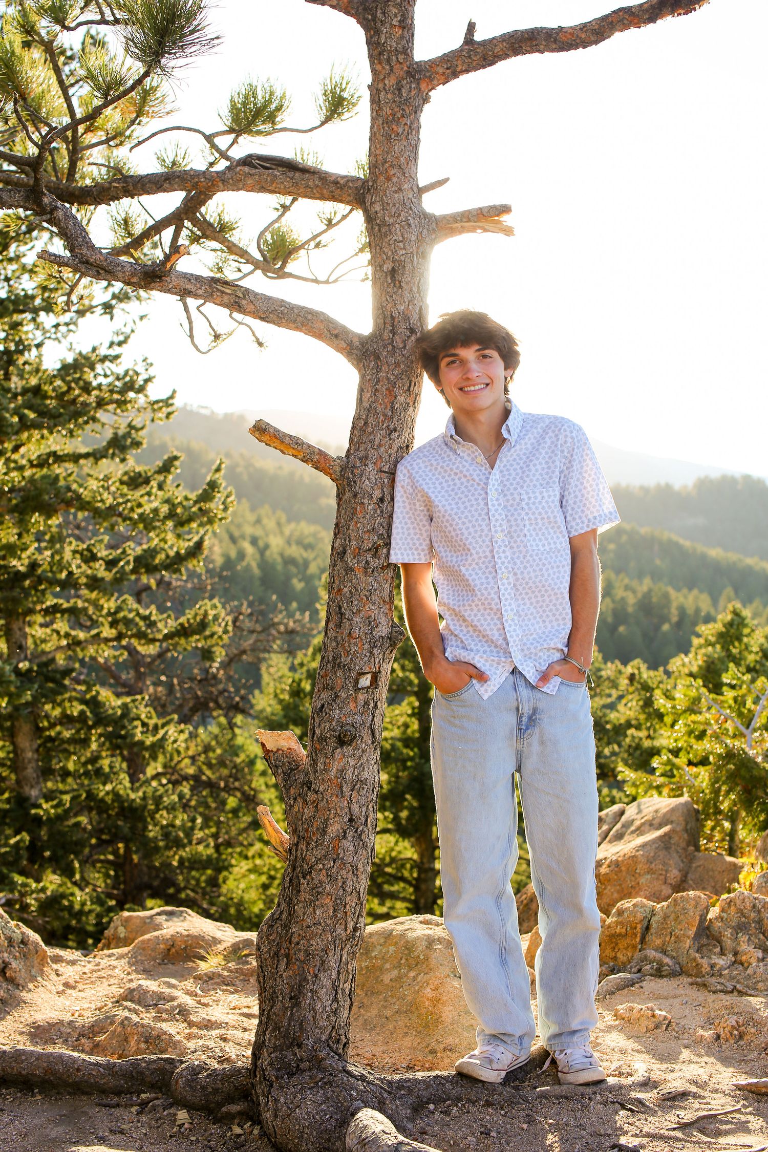 Full-length portrait of someone standing by a pine tree on a rocky mountain overlook wearing casual attire.