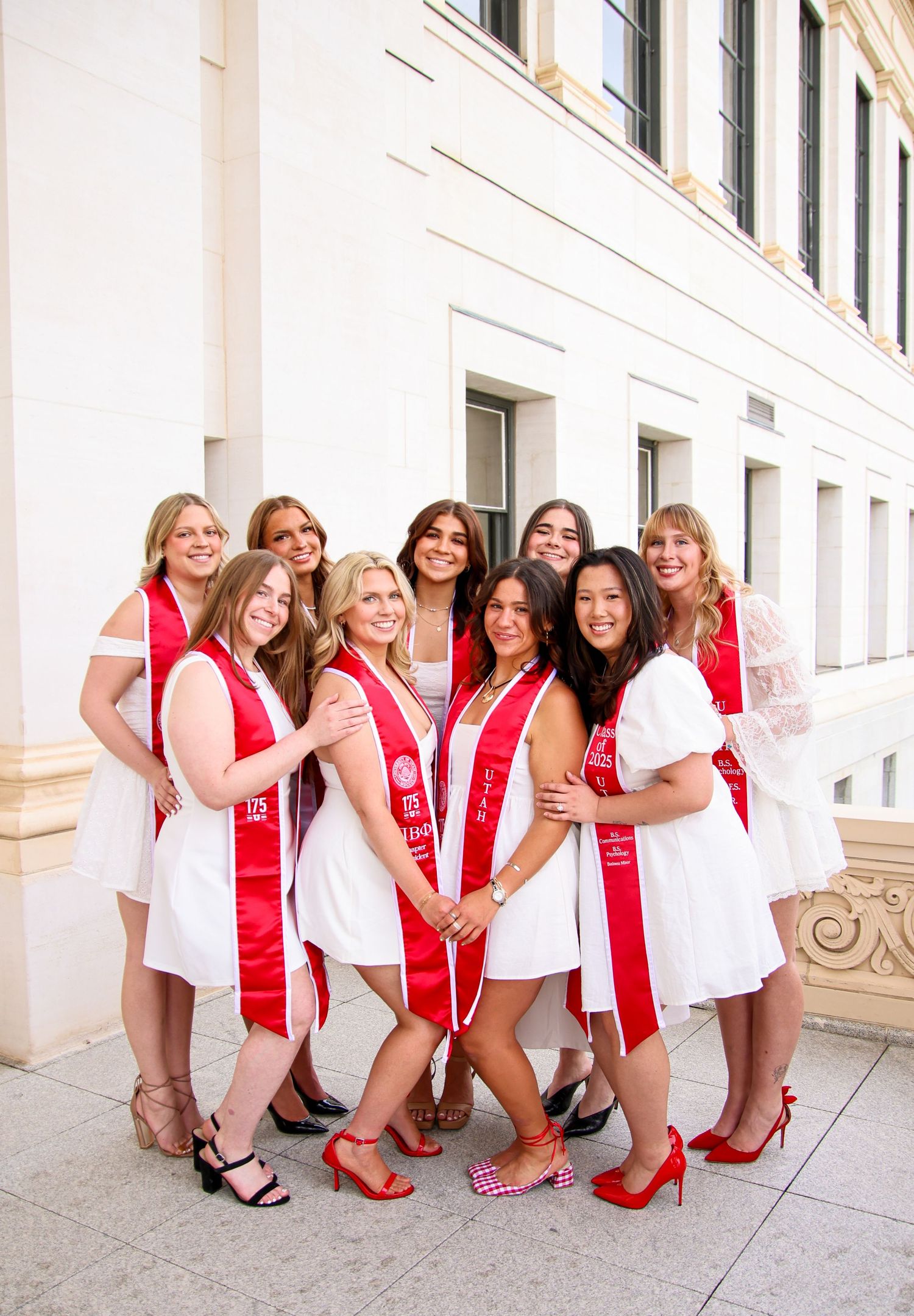 Group wearing matching white dresses and red graduation sashes pose playfully by columns.