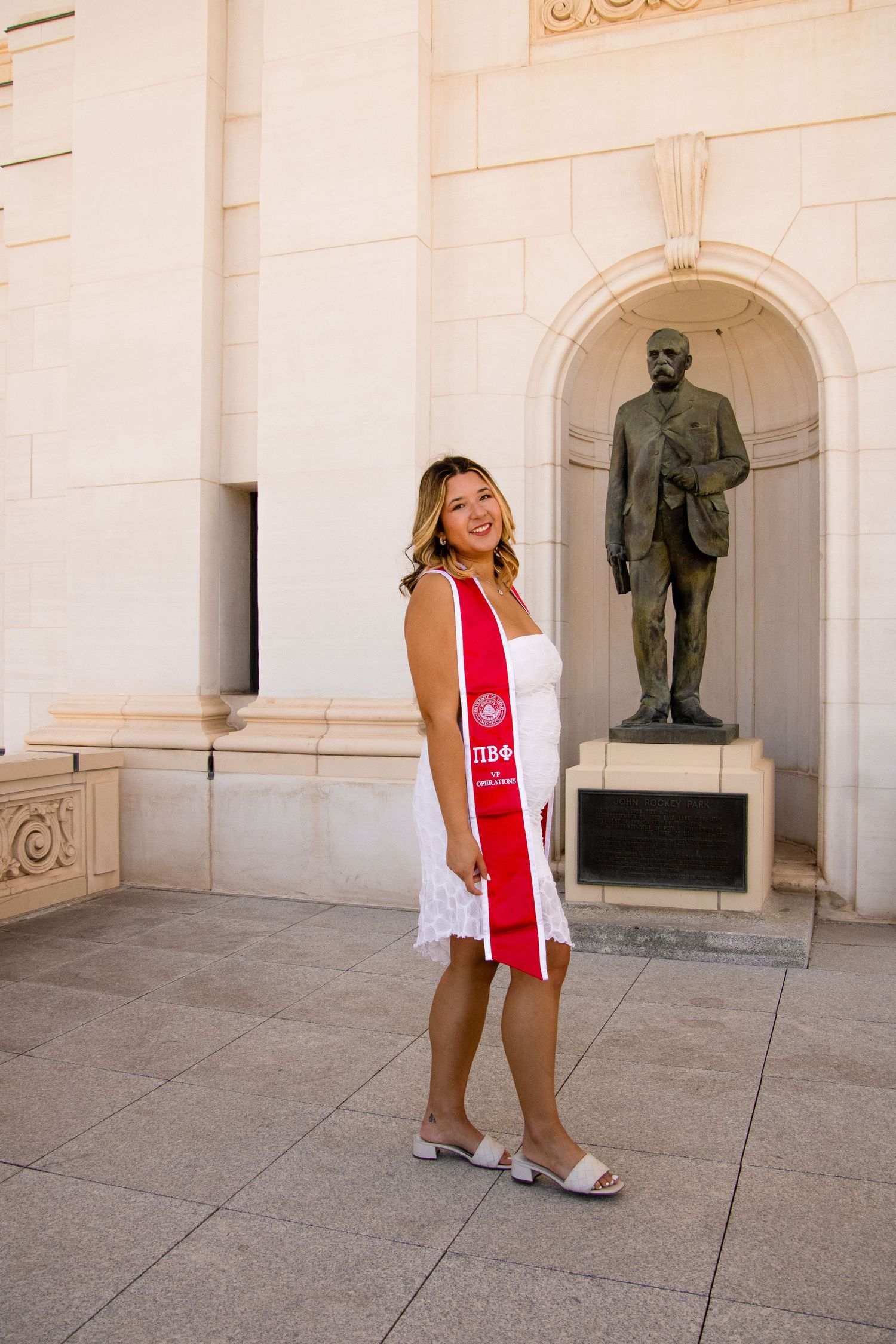 Graduate in white outfit and red Pi Beta Phi stole poses energetically by statue.