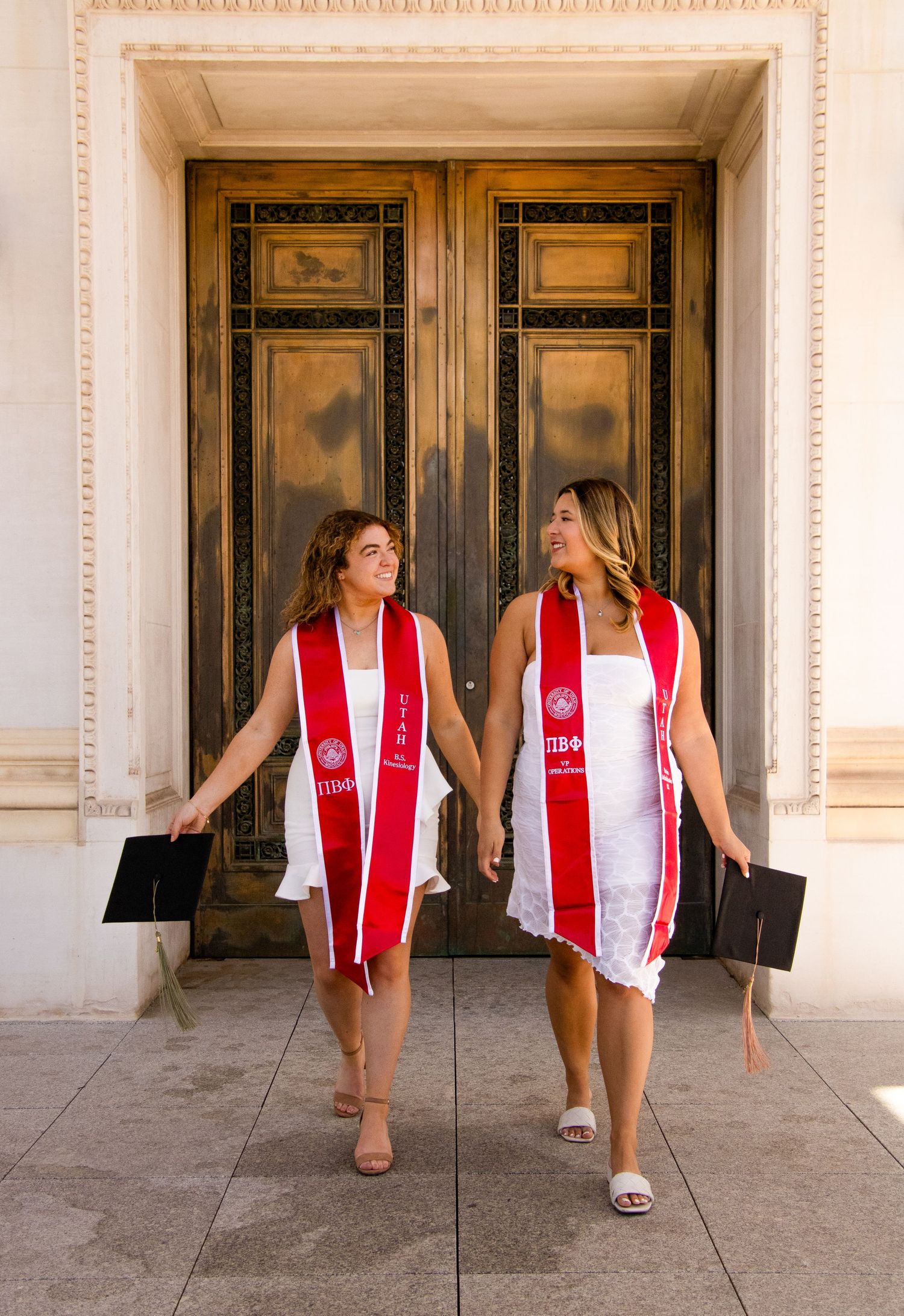 Two graduates in matching red and white stoles pose together by ornate doors.