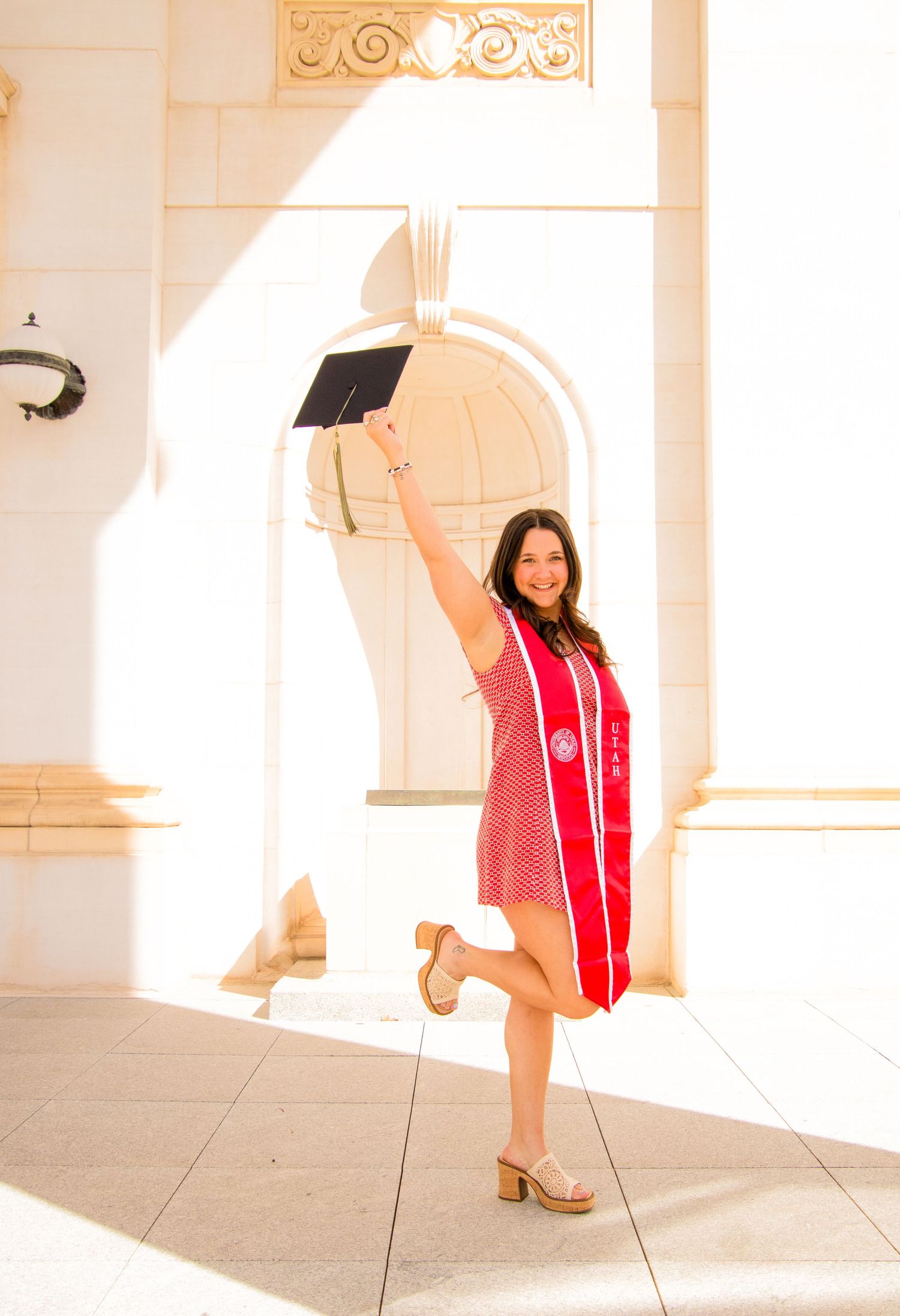 A graduate in red academic regalia poses in various positions along a sunlit architectural walkway.