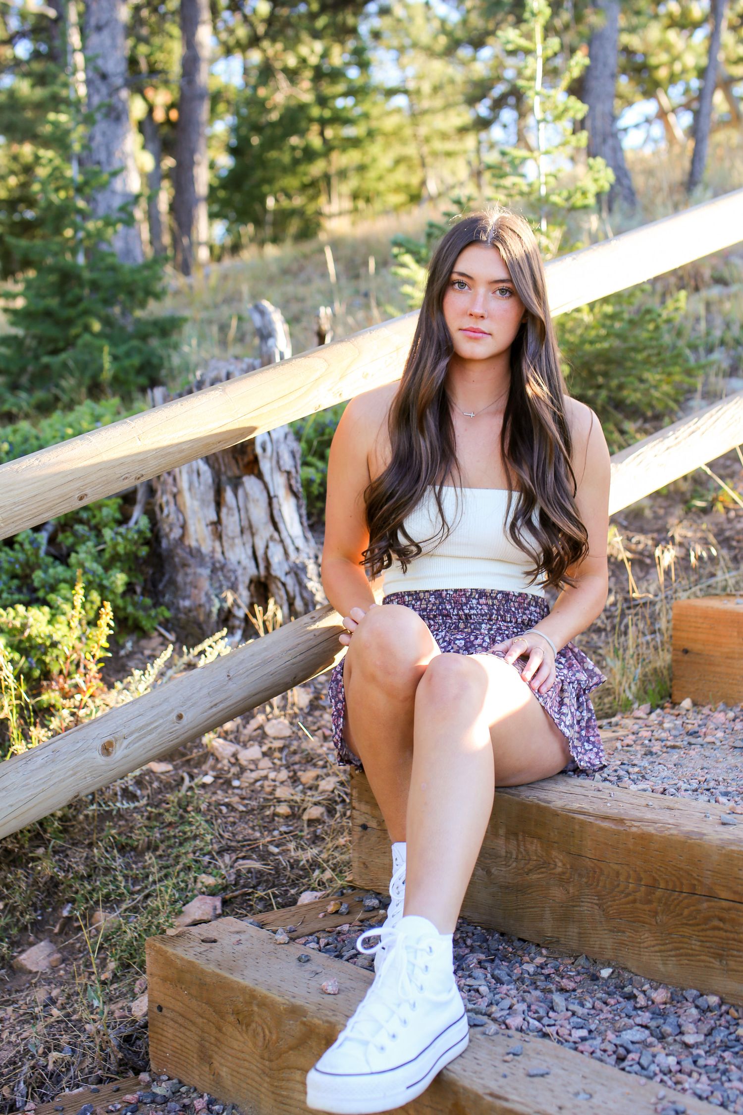 Person in white top and floral skirt sits on wooden steps in forest setting.