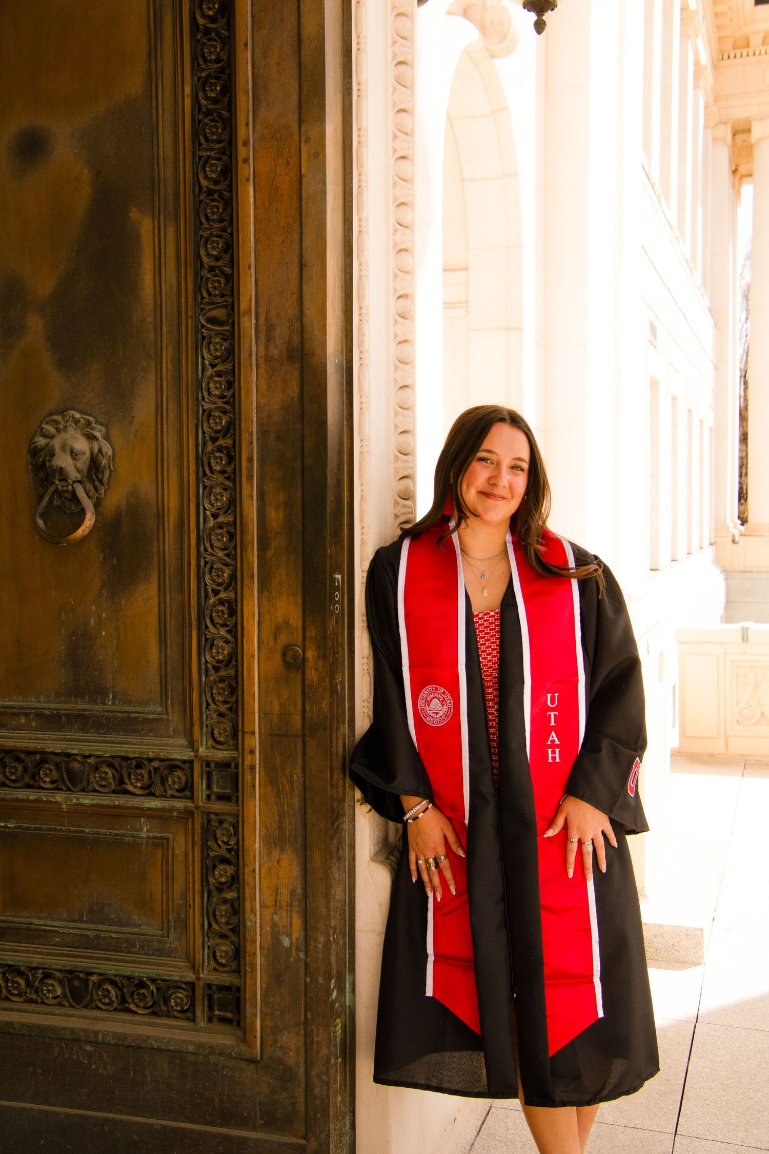 Graduate in black gown and red sash poses by ornate wooden door.
