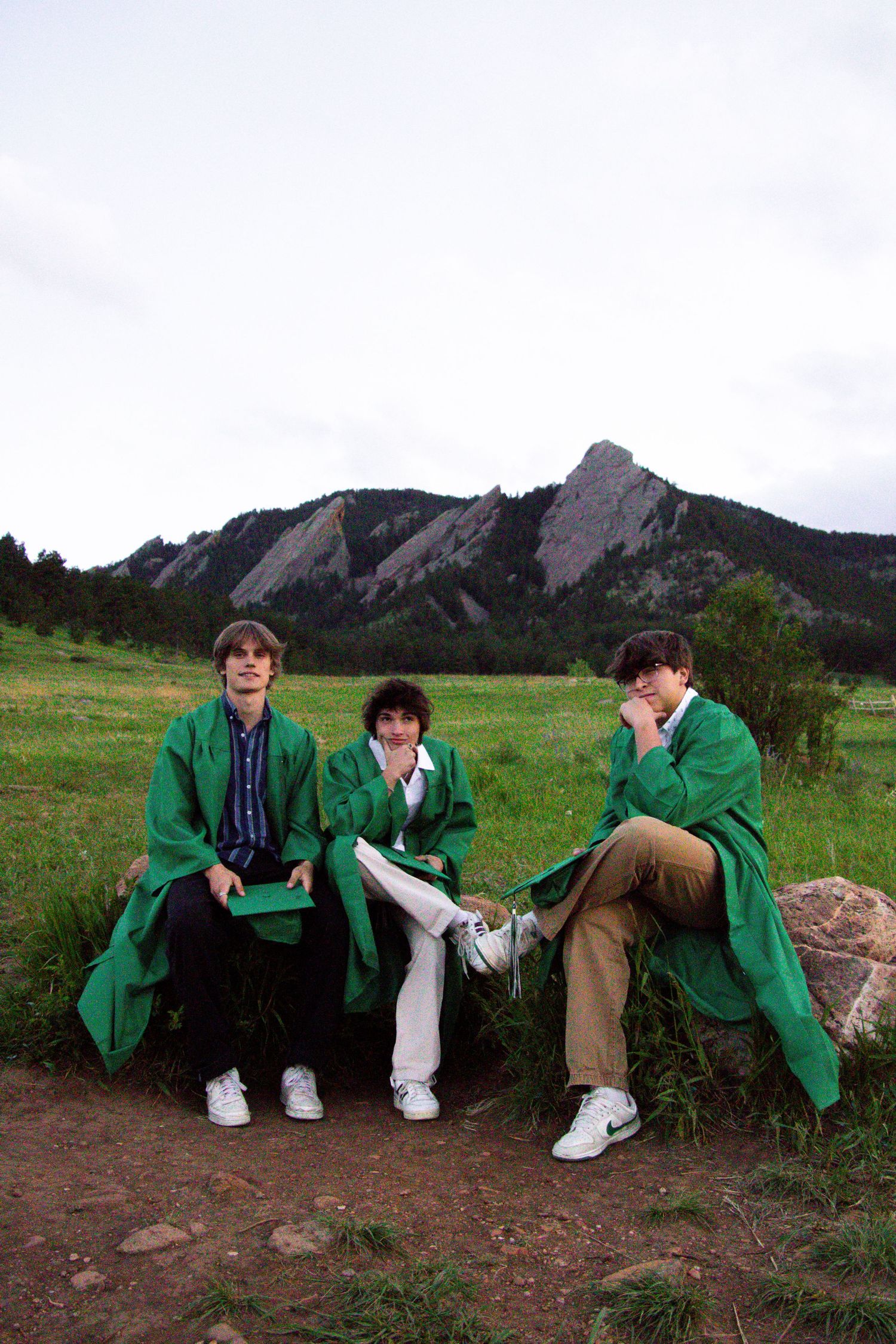 Three graduates in green gowns relax on rocks with the Flatirons mountains in the background.