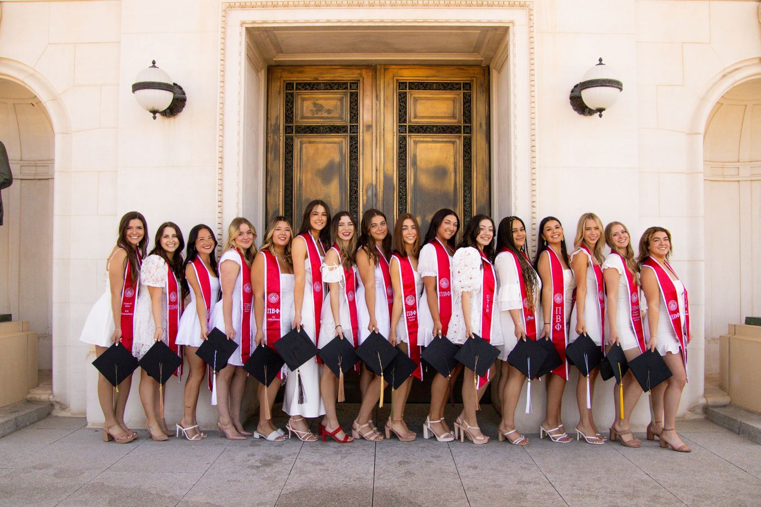 Row of graduates in coordinated outfits pose along classical building exterior.