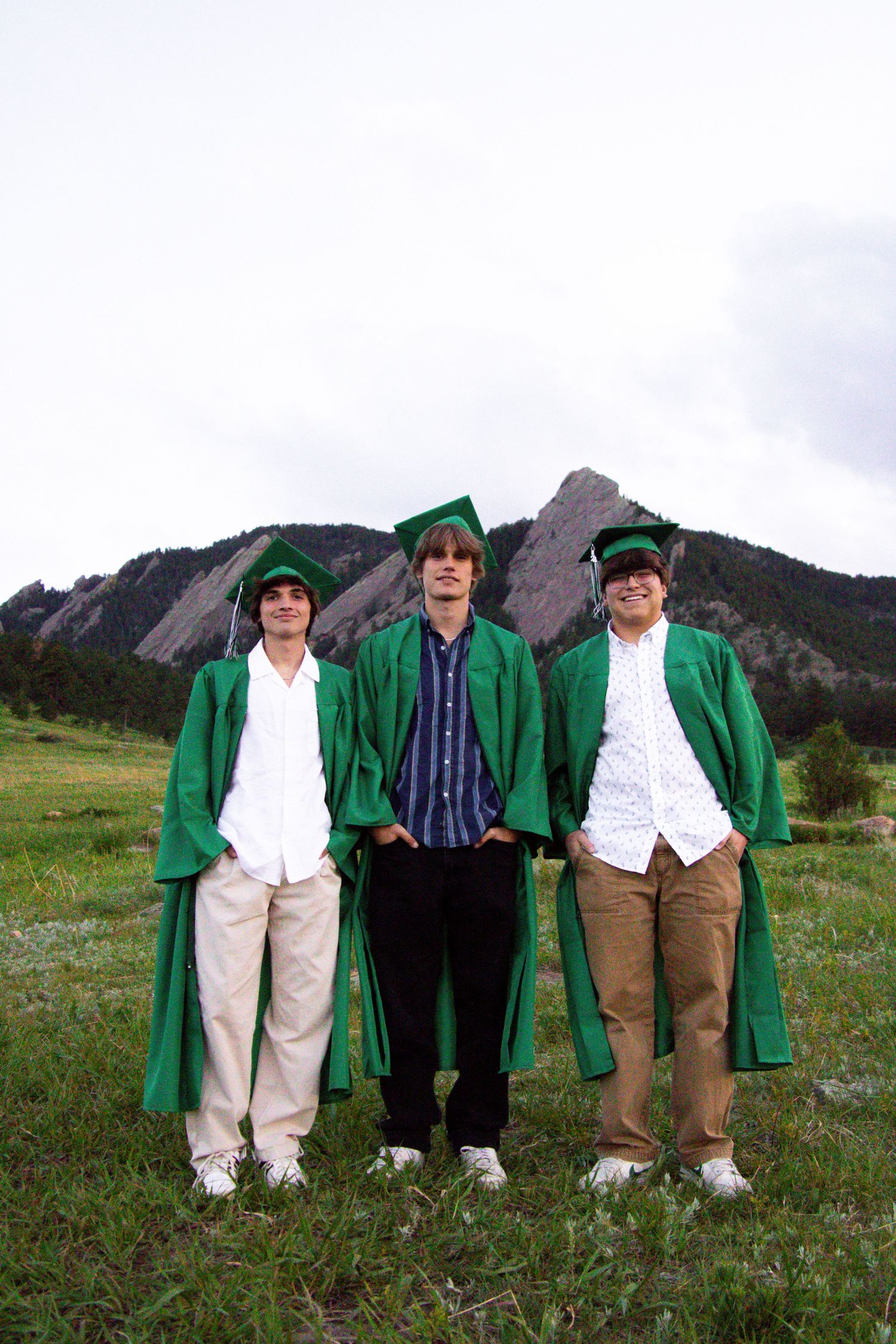 Three graduates wearing green commencement robes pose in a meadow with mountain peaks behind them.