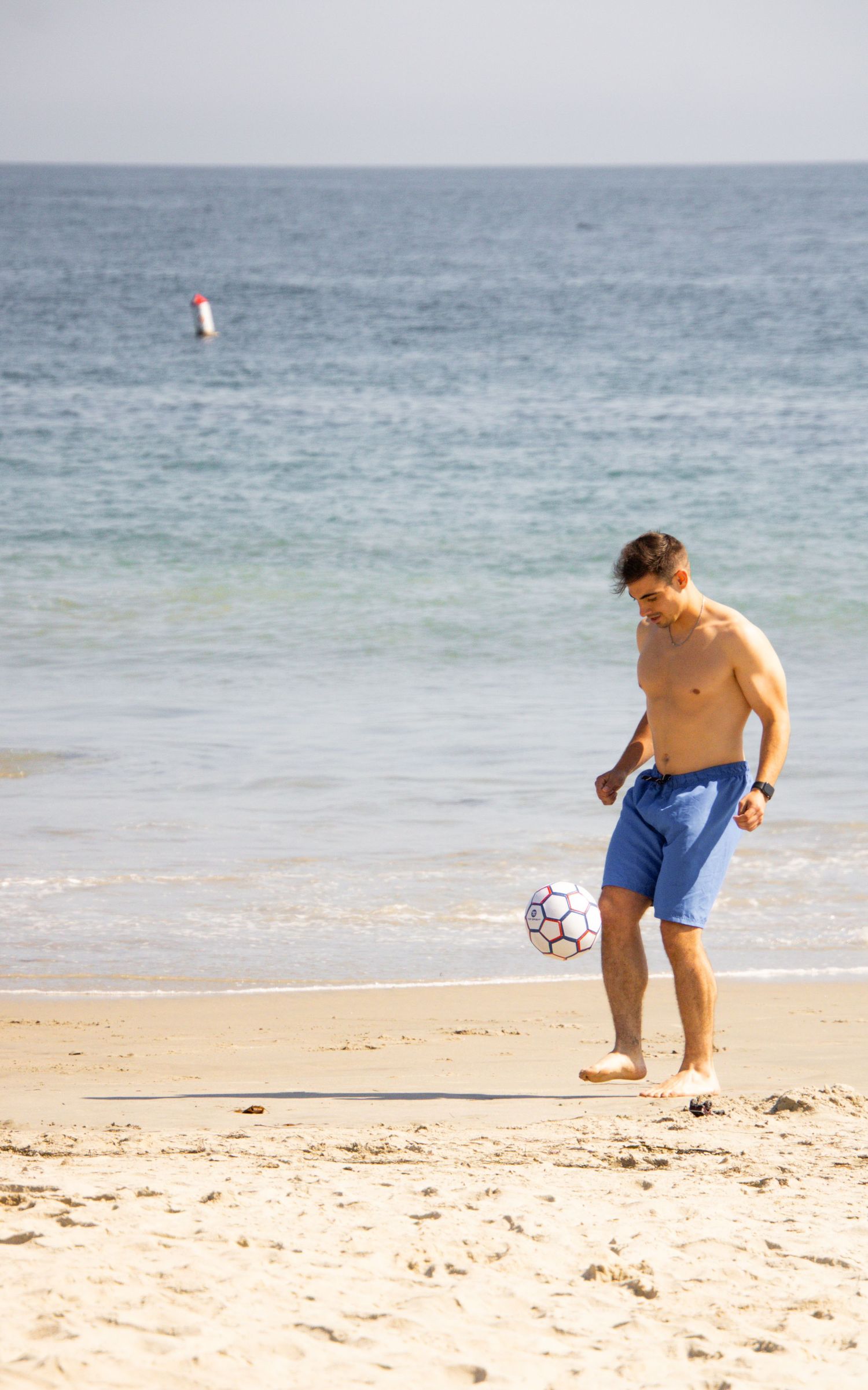 A person in blue shorts plays soccer on a sunny beach with ocean waves in the background.