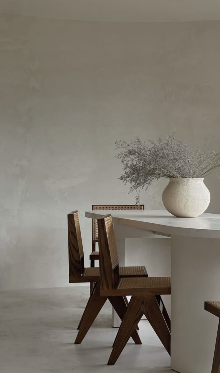 Minimalist dining setup with wooden angular chairs and white round vase containing dried flowers on concrete table.