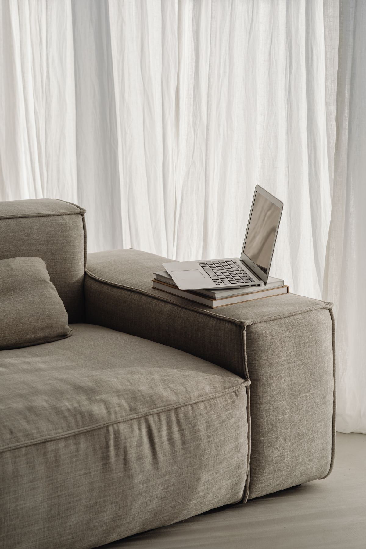 Minimalist gray modular sofa section with laptop against white curtained backdrop in modern living space.