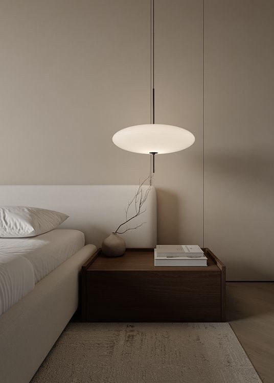 A minimalist bedroom interior with neutral tones features a low platform bed and simple pendant light against beige walls.