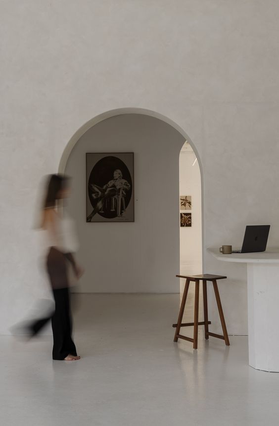 Motion-blurred figure walks through minimalist white room with arched doorways and wooden furniture against bare walls.