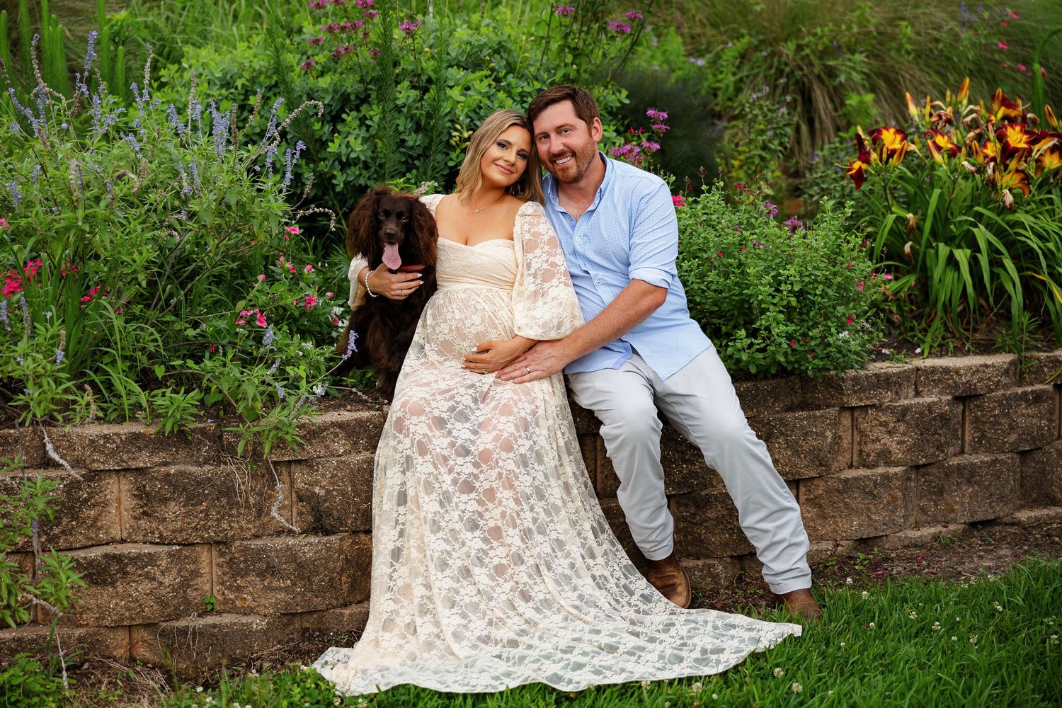 Expecting couple posed on low wall, surrounded by flowers and hugging their dog for a portrait by Memphis photographer Emily Joy Photograpy