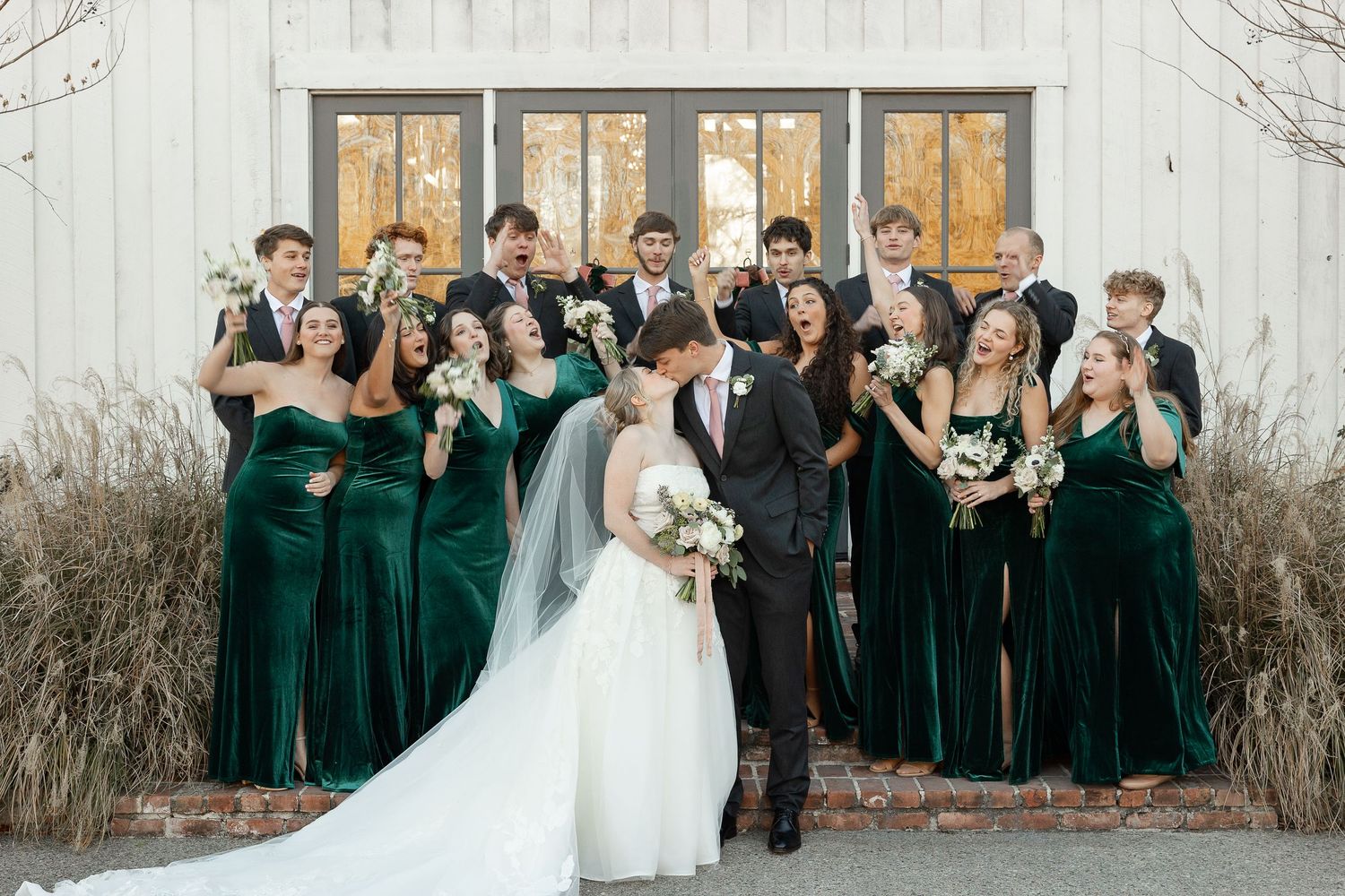 Eight bridesmaid and eight groomsmen cheer as a bride and groom kiss in front of the Robinshaw wedding venue.
