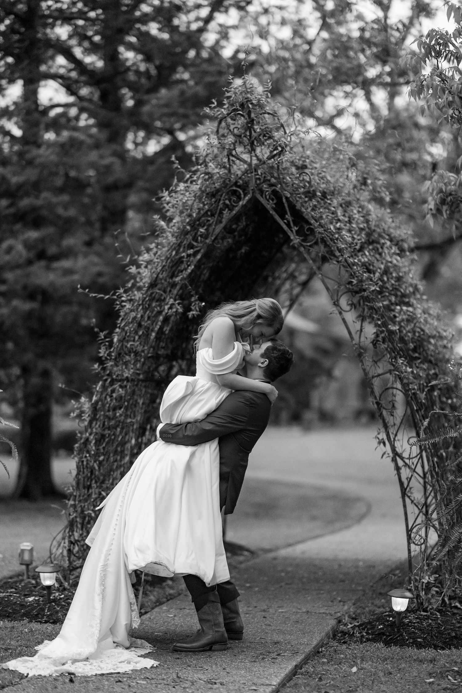 A groom lifts his bride for a kiss under a garden arch. Image by Emily Joy, wedding photographer in Memphis.