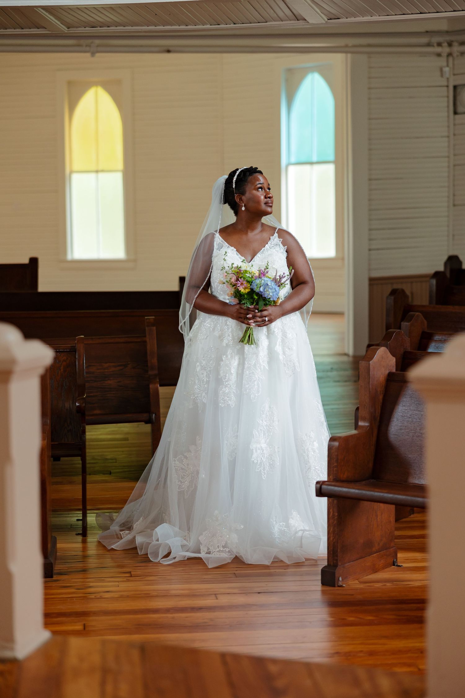 A bride in an elegant gown stands in a chapel with stained glass windows for portrait by Memphis Wedding Photographer Emily Joy Photography