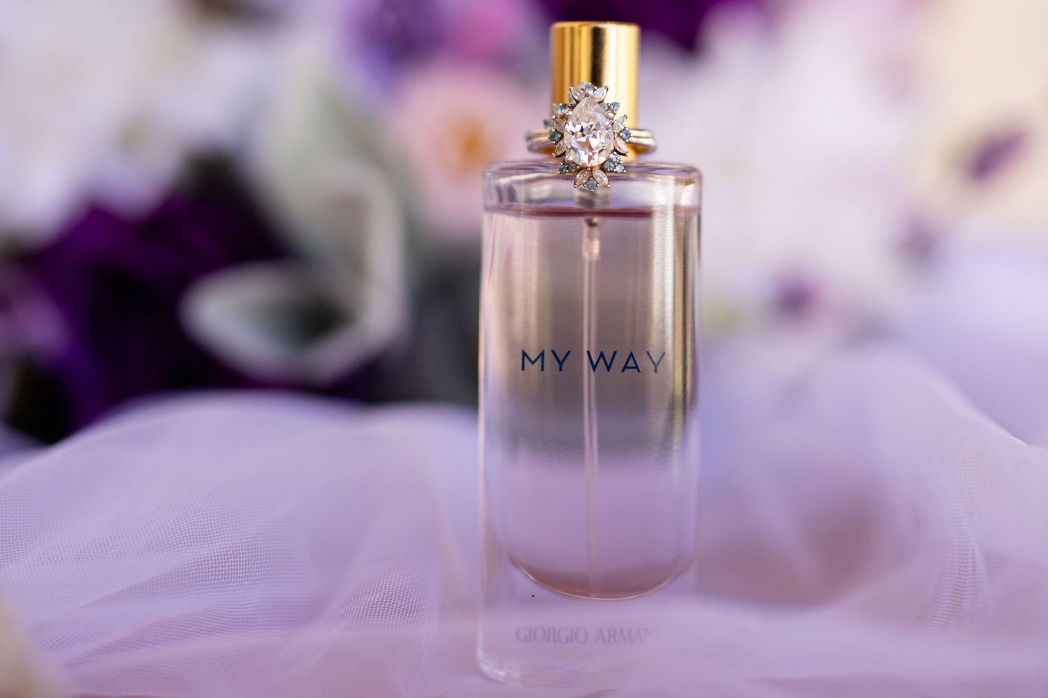 A bride's engagement ring displayed on her perfume bottle with tulle and a bouquet in the background