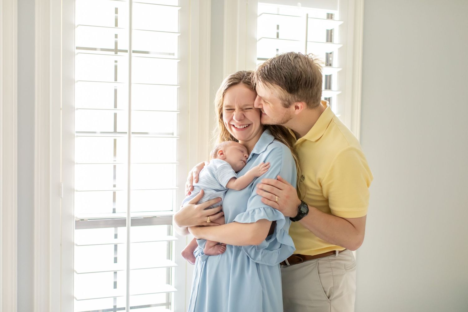 A husband kisses his wife as she holds their newborn son for a portrait by Emily Joy Photography, now a weddinggrapher Emily Joy Photography