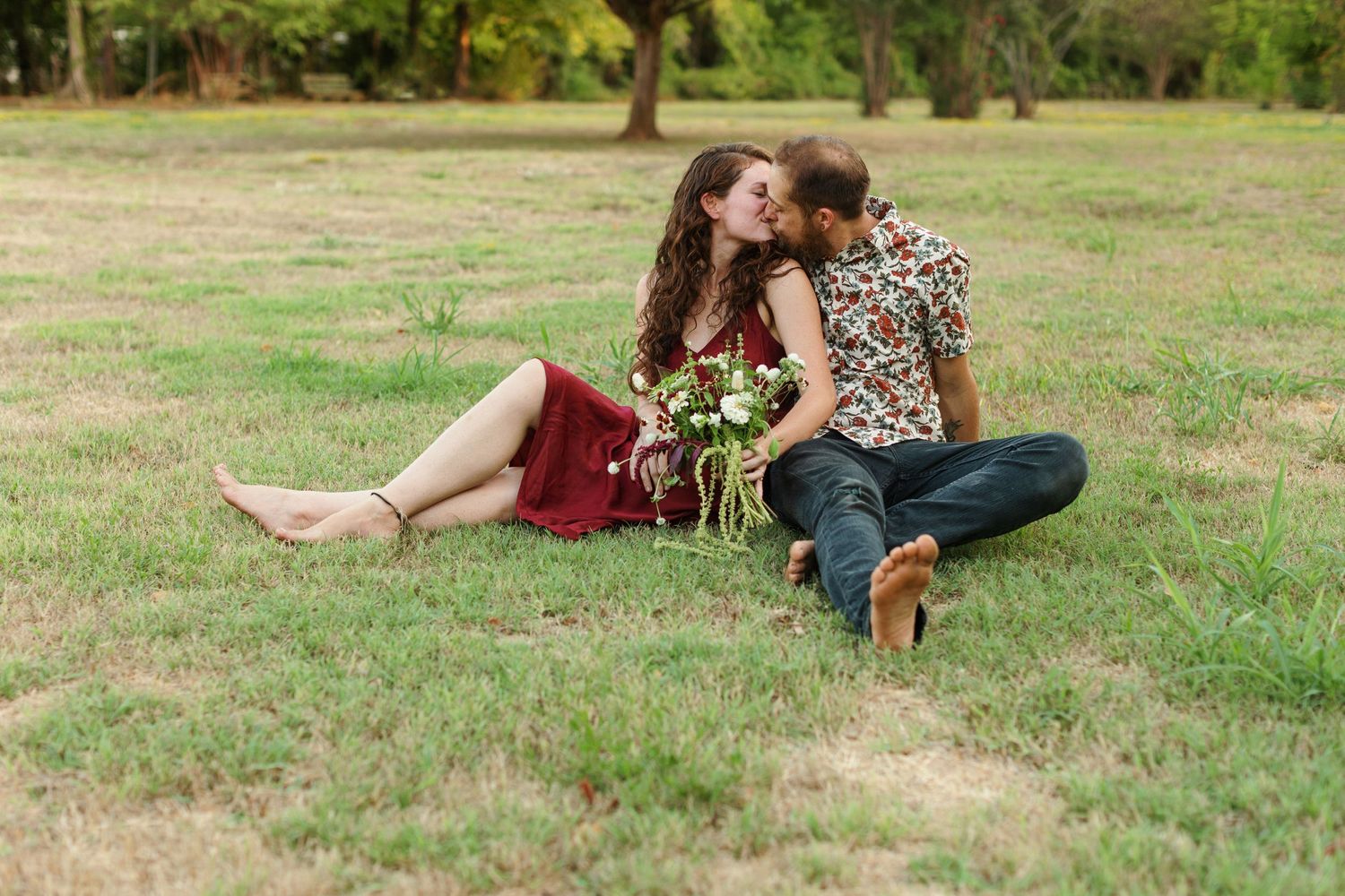 Engaged couple, sitting in the grass, shares a kiss for a portrait  by Emily Joy Photography, now a wedding photographer in Memphis, TN.