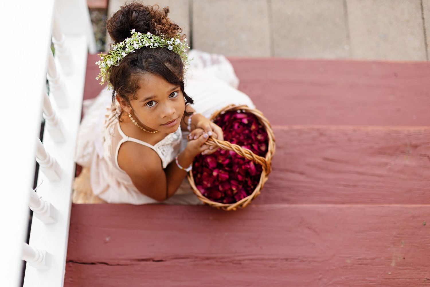 A flower girl smiles up at the camera while she and holds a basket of bright pink rose petals photographed by Emily Joy Photography.