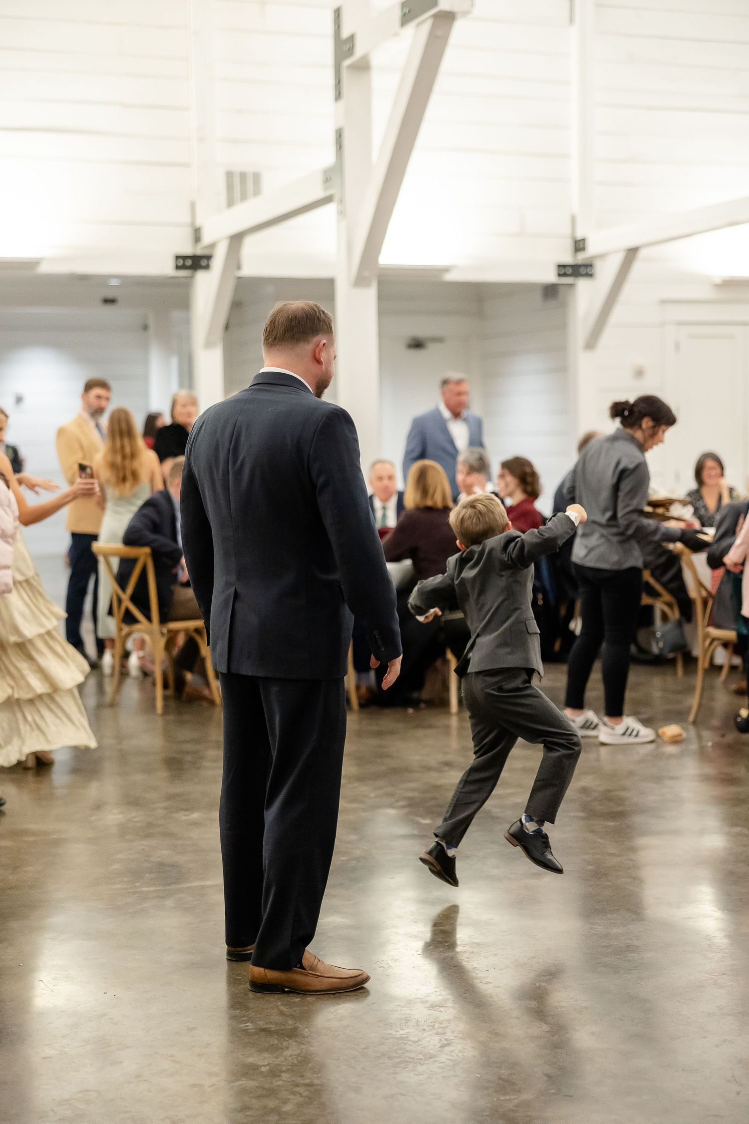 A young boy in a dress suit dances energetically and is pictured here in mid-air while his father watches.