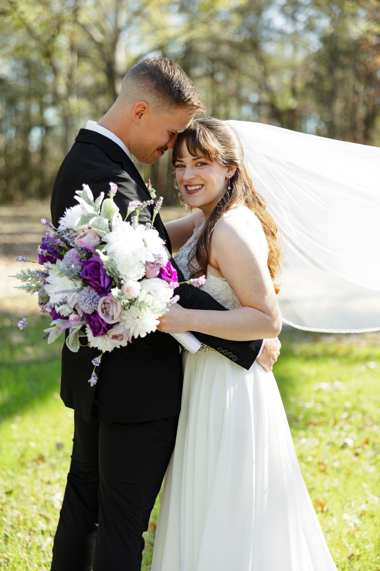 A groom leans toward his bride while she smiles at the camera. Her veil streams behind her.