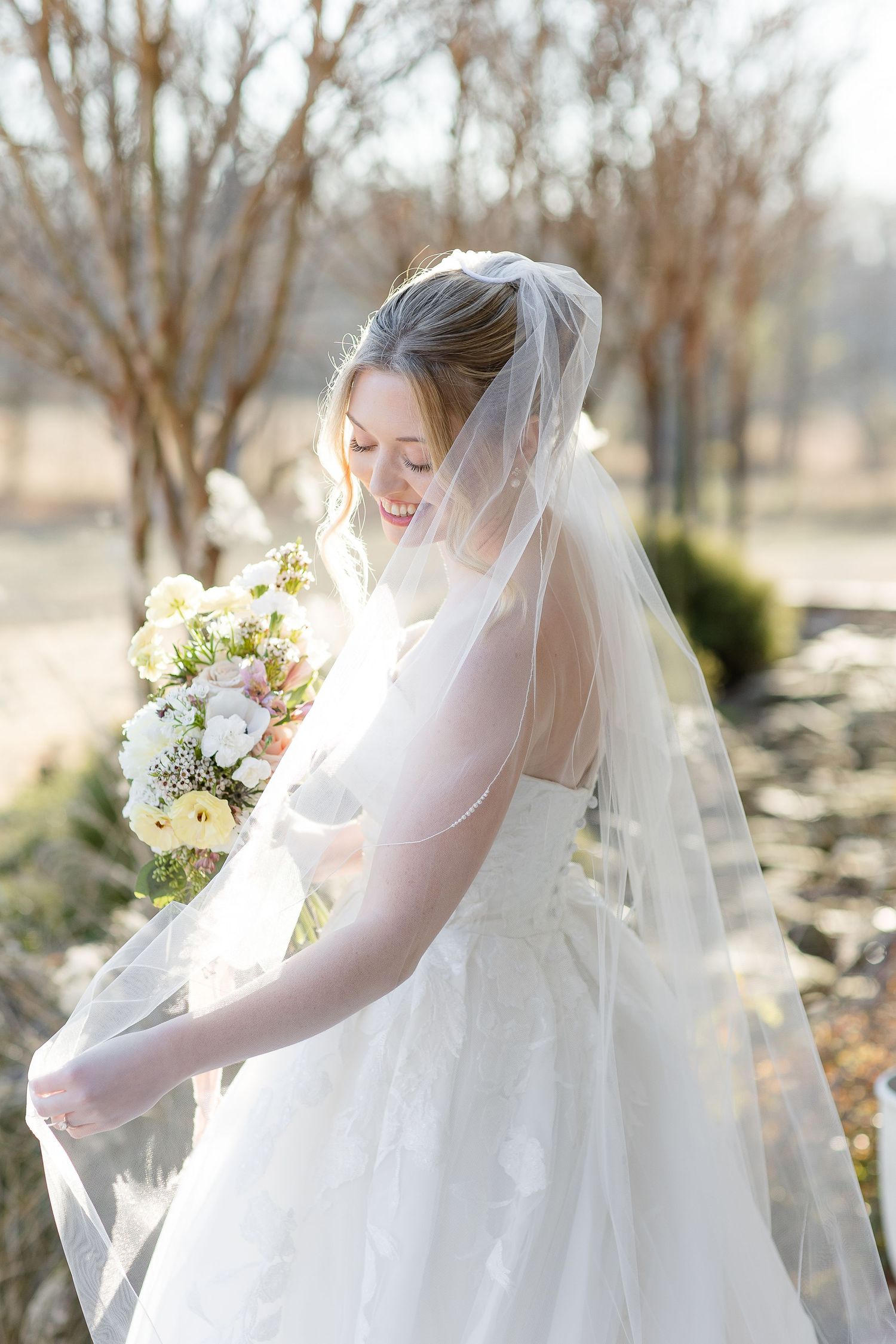 An elegant bride fluffs her veil, while smiling gently outside on a sunny day. Image by Emily Joy, wedding photographer in Memphis.