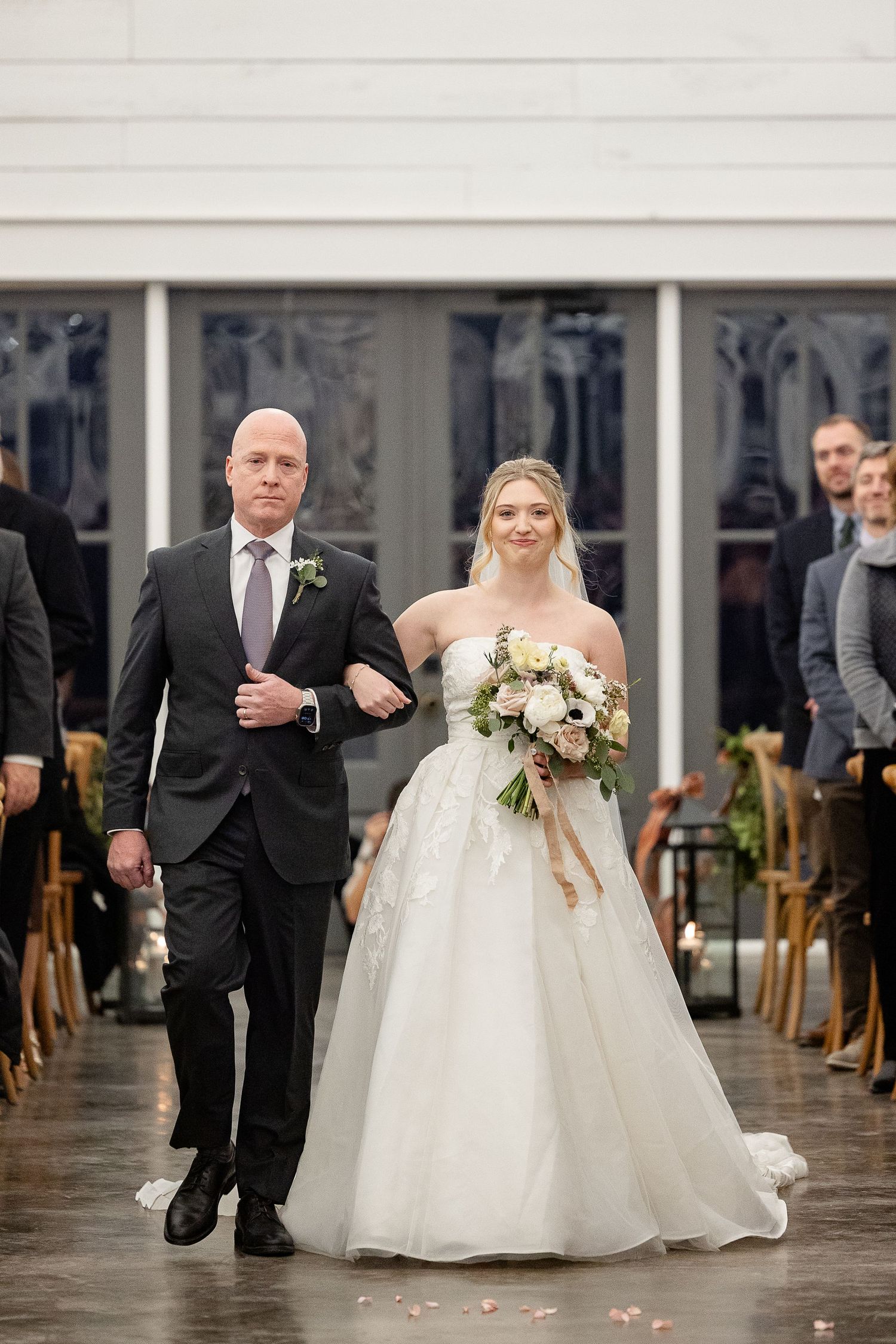 A bride in a strapless white dress with a lace overlay is escorted down the aisle by her father at the Robinshaw wedding venue.