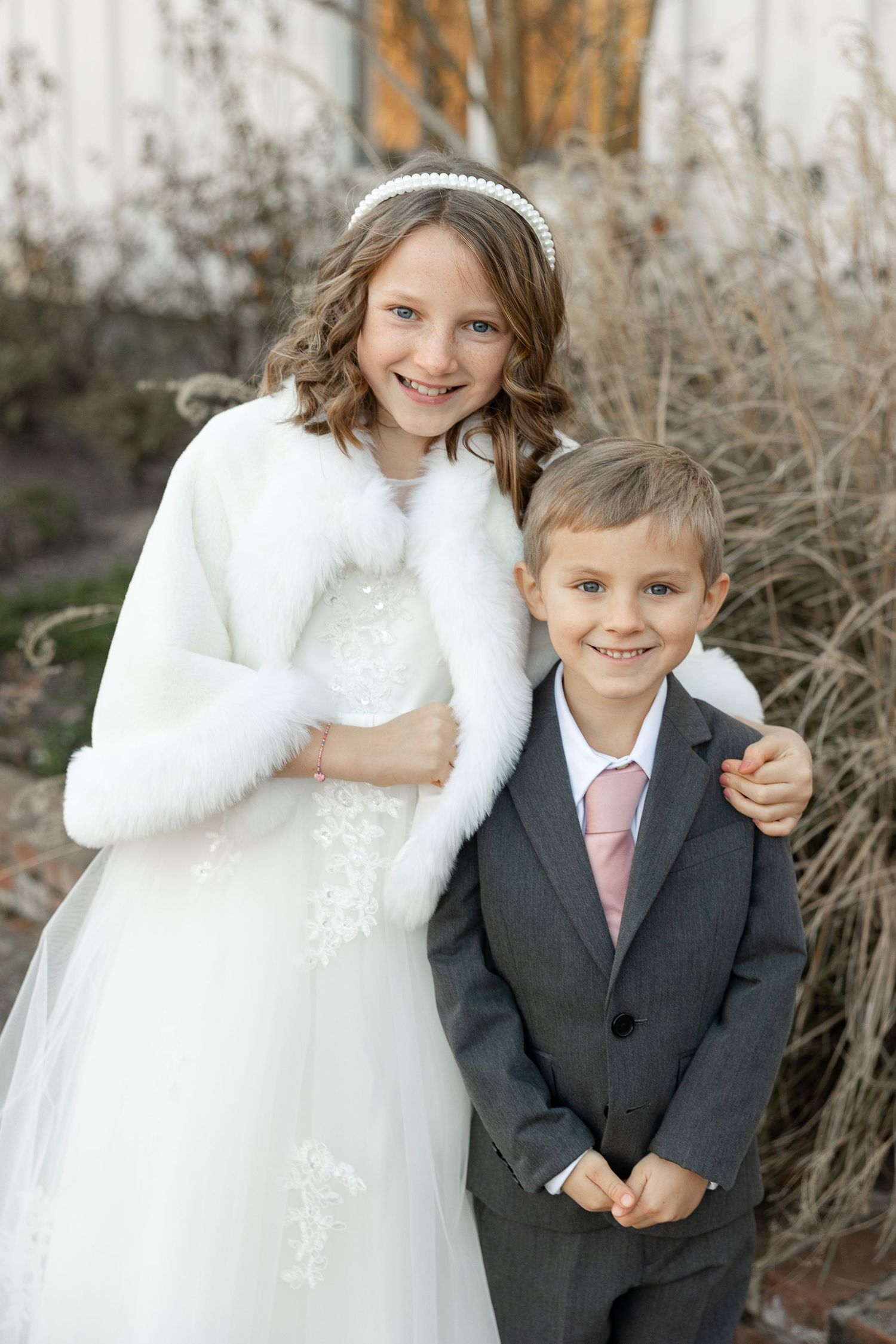 A young brother in a suit and sister in a white dress with a fur shawl smile at the camera for a portrait as flower girl and ring-bearer.