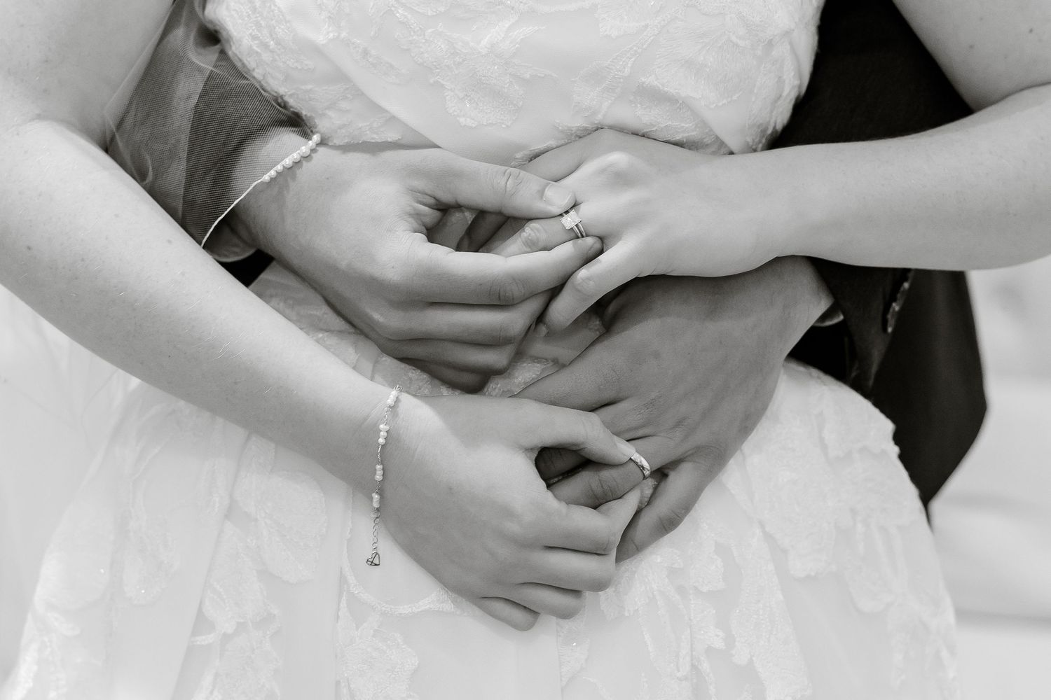 A groom hugs his bride from behind and they hold each other's hands and display each other's wedding rings.