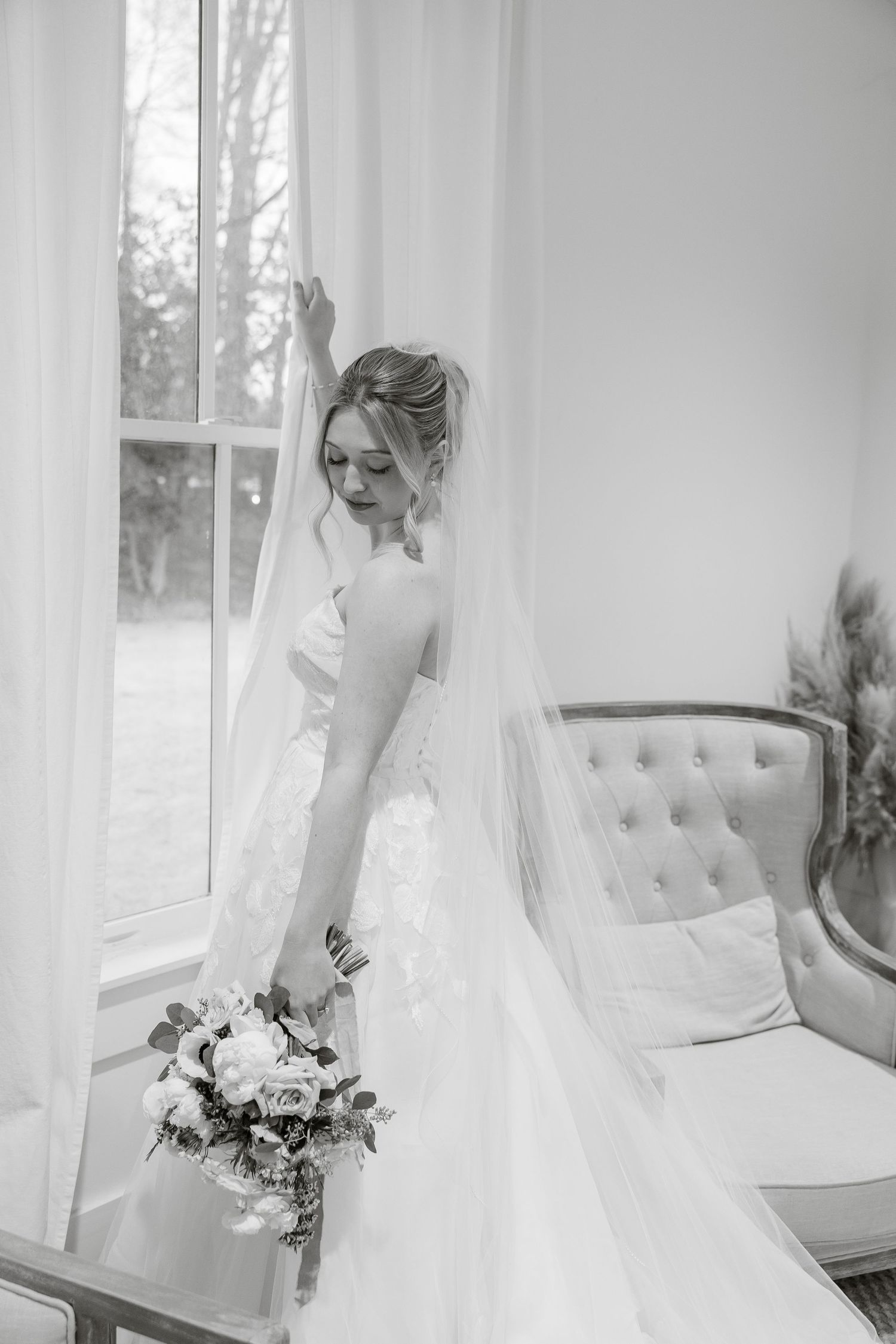 A bride holds curtains at window for a black-and-white editorial-style portrait by Memphis wedding photographer, Emily Joy Photogpraphy.