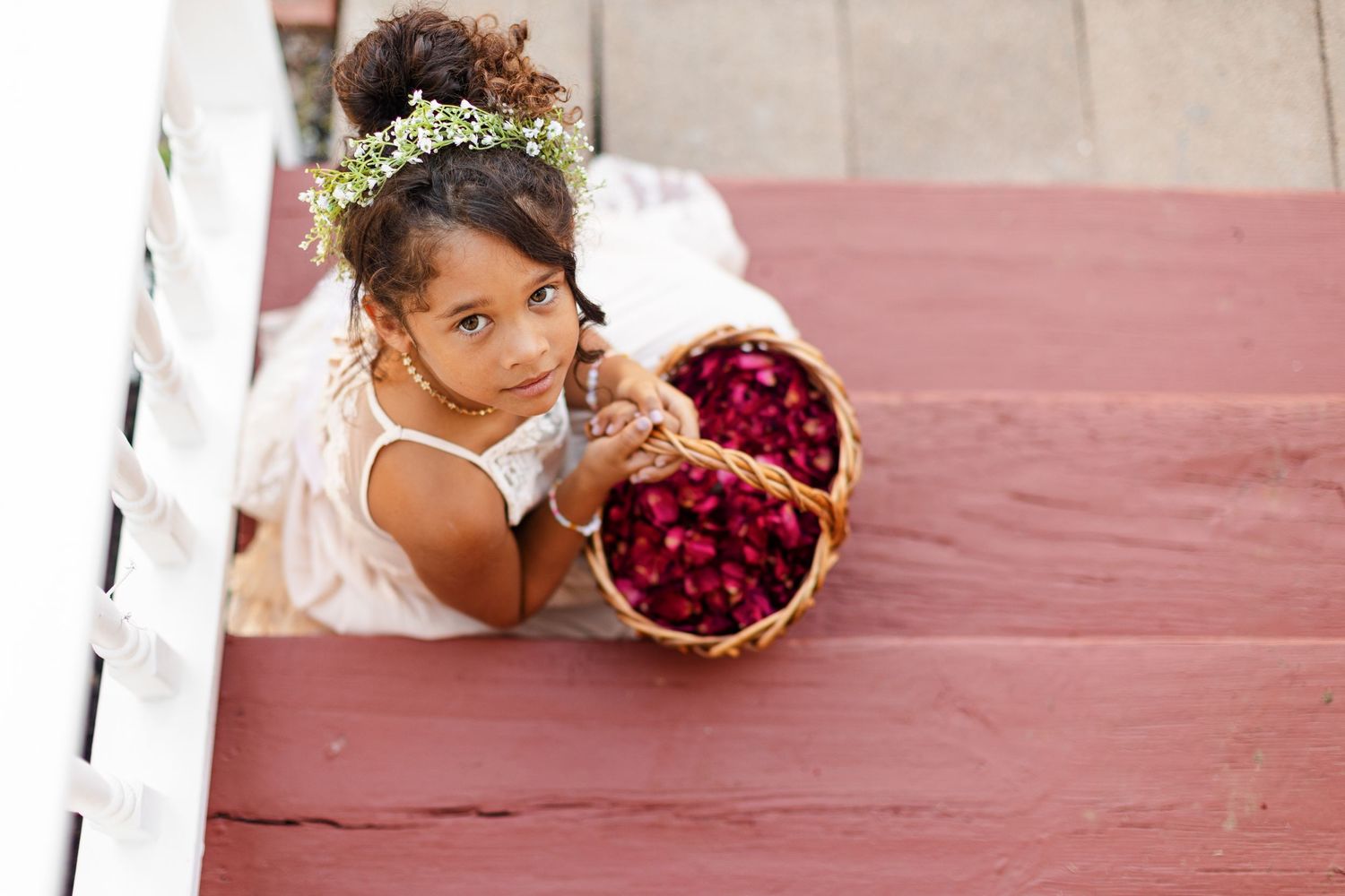 A flower girl holding a basket of pint rose petals smiles gentle up at the camera. Image by Emily Joy, wedding photographer in Memphis.