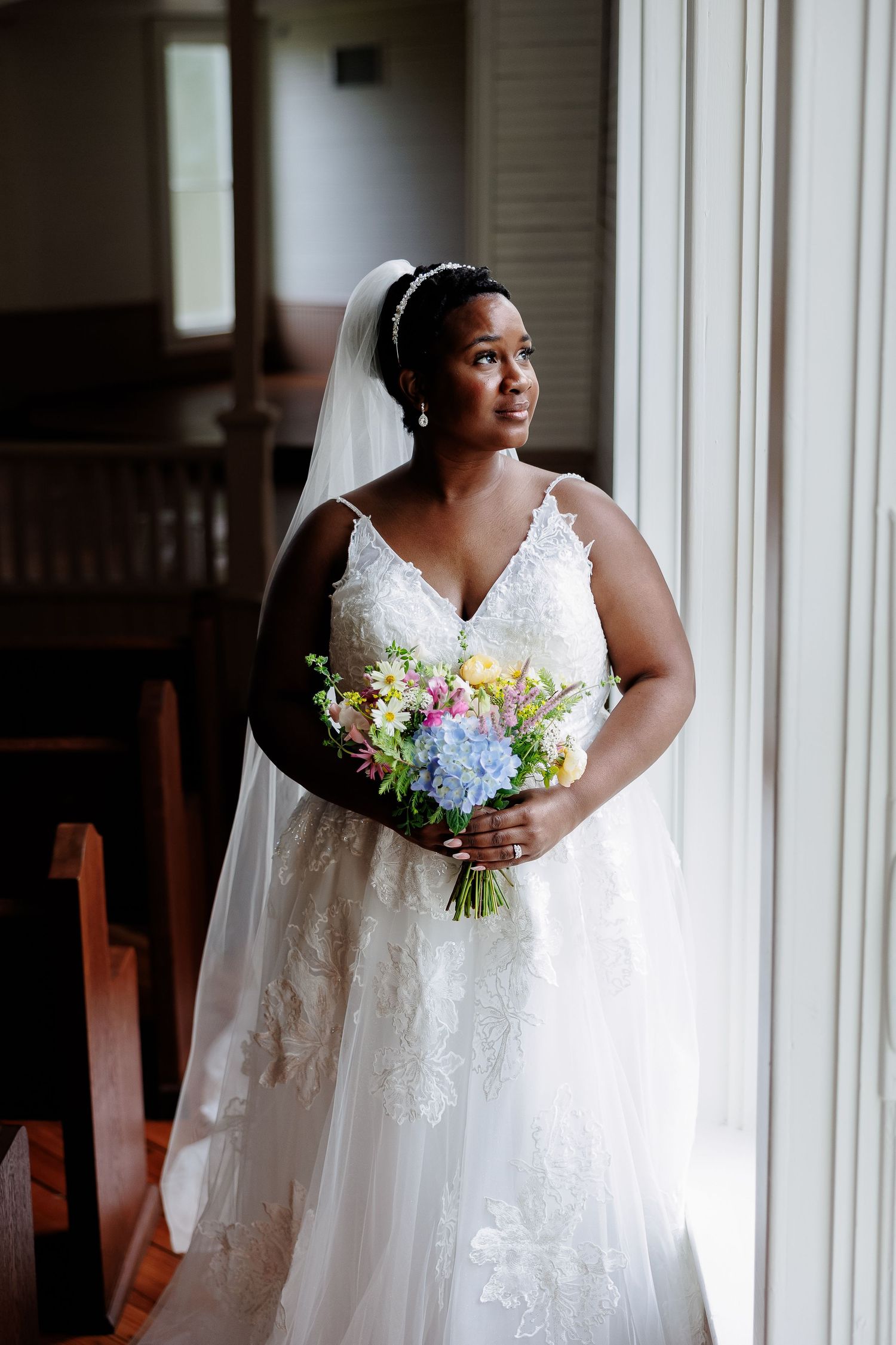 An elegant bride holding a pastel bouquet looks out a window glamorously. Image by Emily Joy, wedding photographer in Memphis.