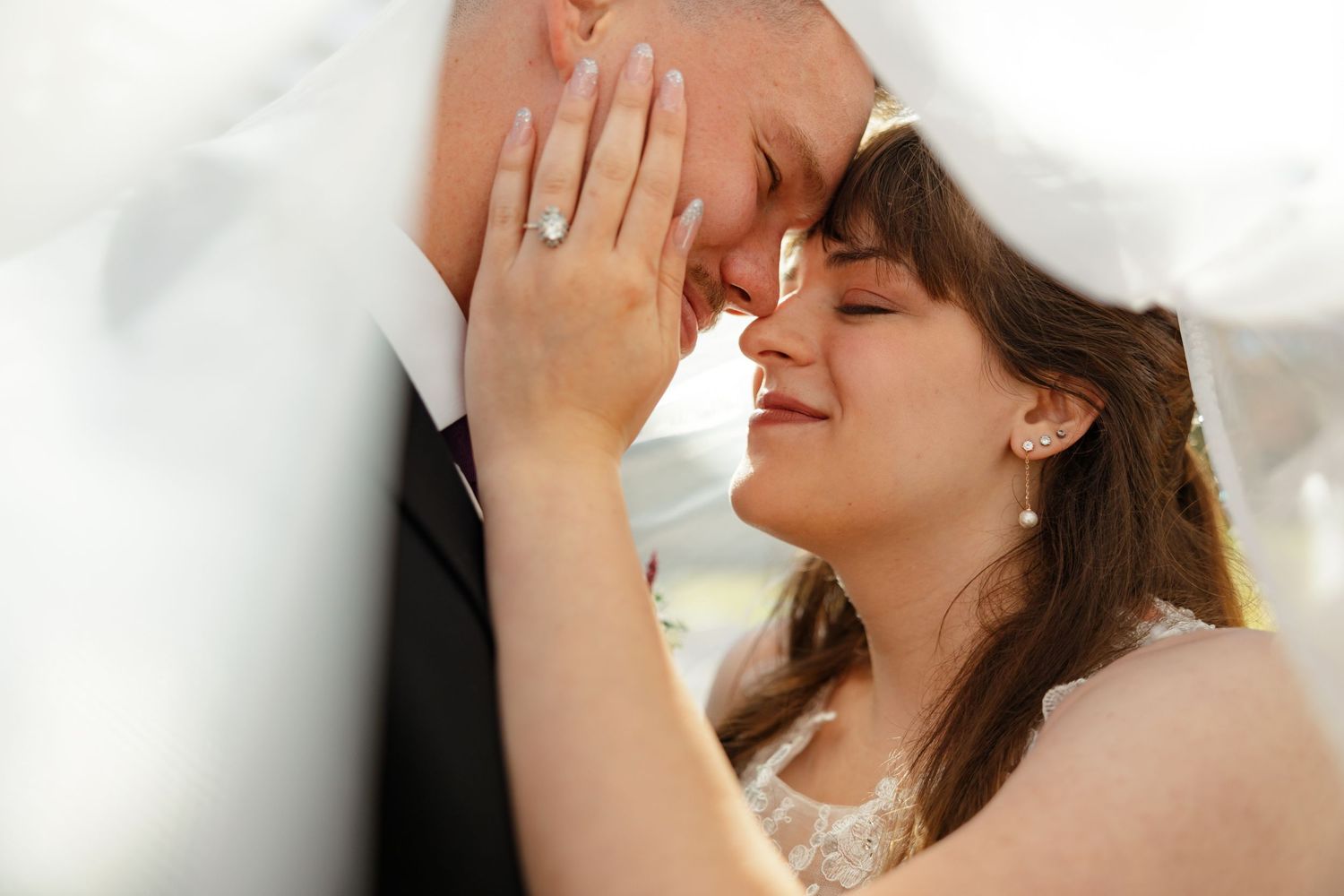 A bride and groom smile gently while the bride caresses the grooms face, revealing her engagement ring.