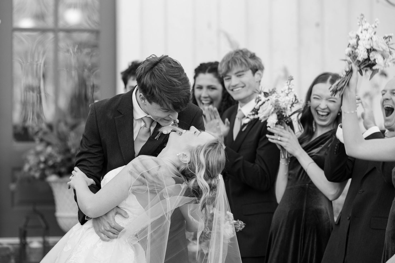 A bride and groom laugh after a dip-kiss while wedding party members look on and cheer.
