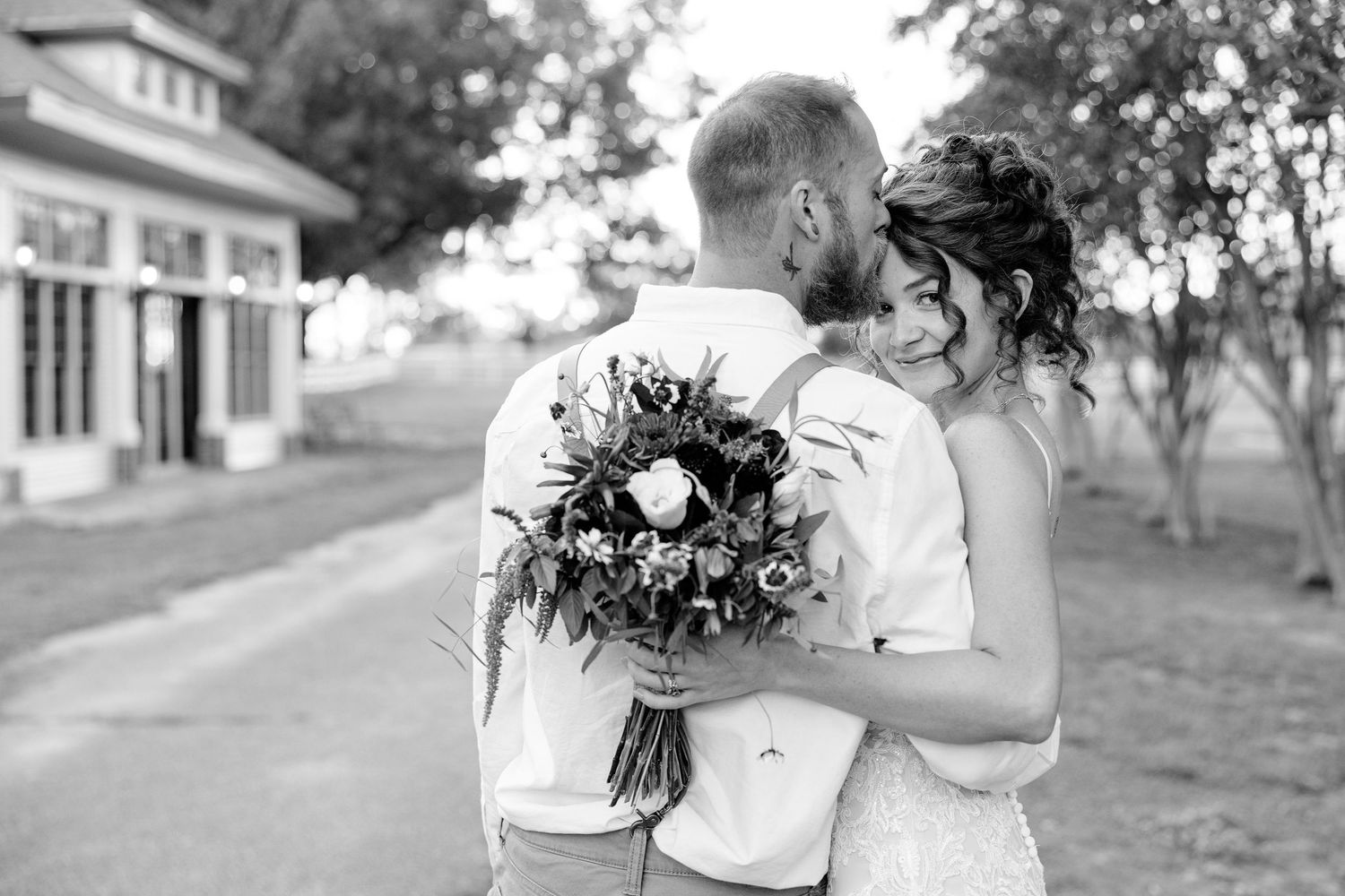 A groom kisses his bride on the forehead while she smiles at the camera. Image by Emily Joy, wedding photographer in Memphis.