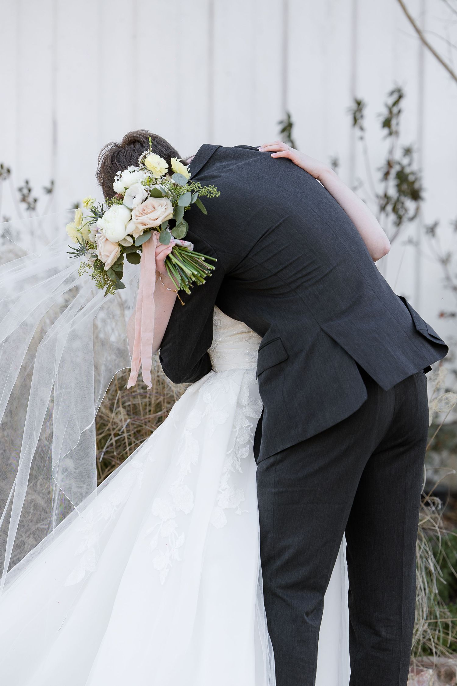 A groom hugs his bride while the wind blows and she holds her bouquet.  Taken outside the Robinshaw wedding venue.