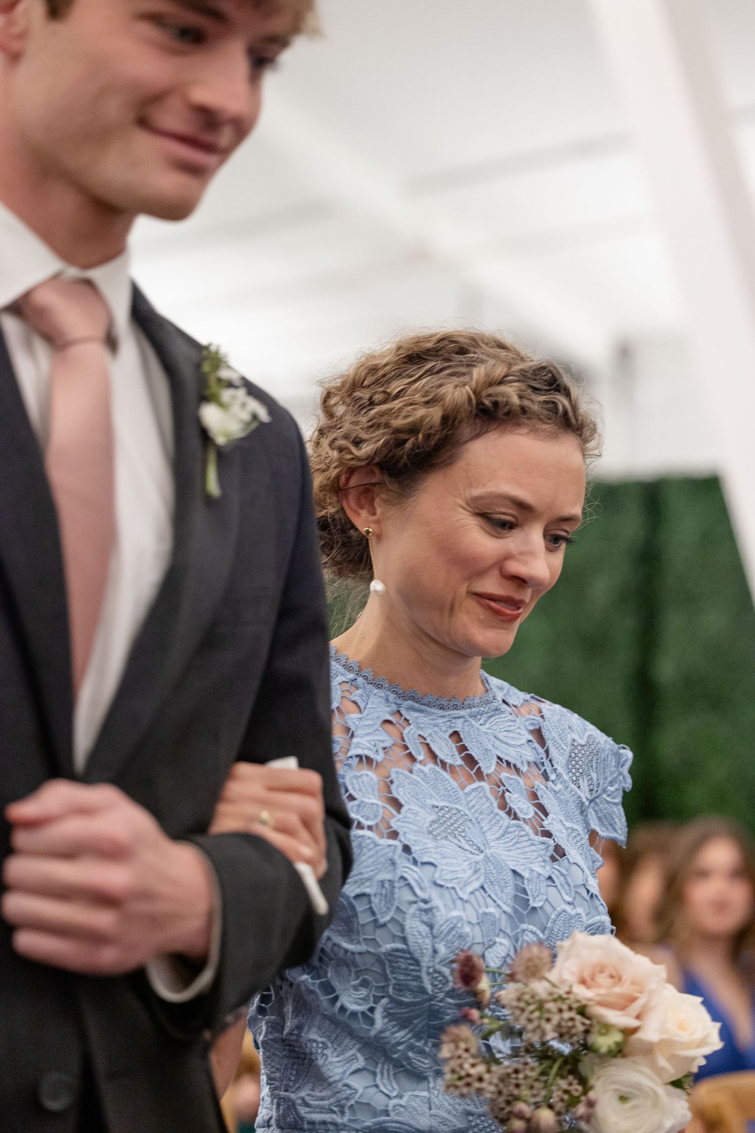 The mother of the bride in a light blue lace dress smiles gently as her son escorts her to her seat.