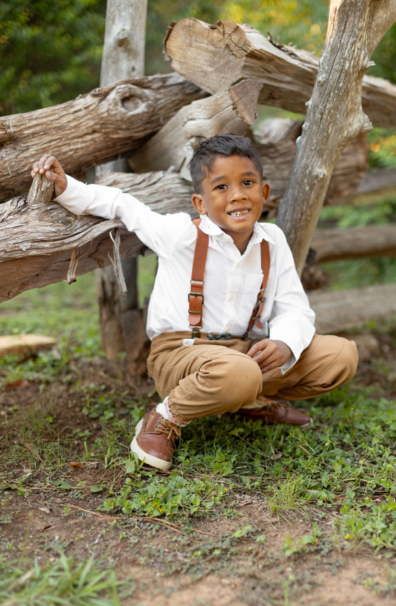 A cute young ring bearer in suspenders crouches down for a portrait by a rustic wooden fence.