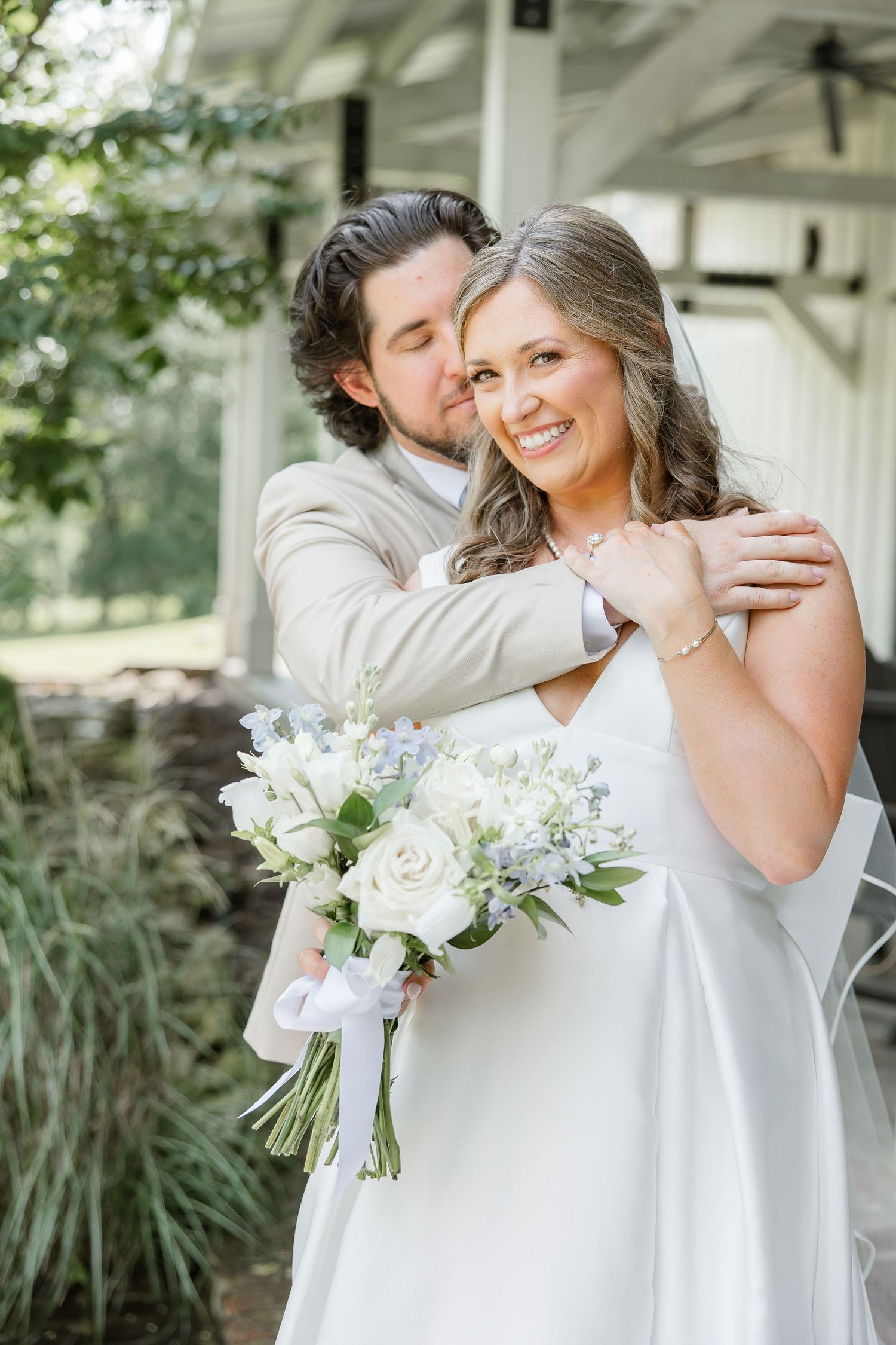 A groom cuddles his bride while she smiles, holding her bouquet. Image by Emily Joy, wedding photographer in Memphis.