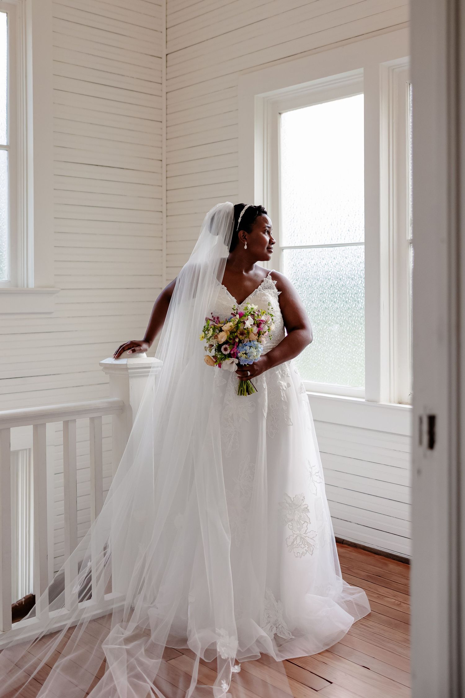 A bride in an elegant white wedding gown looks out a bright window for a portrait by Emily Joy, a wedding photographer in Memphis, TN.