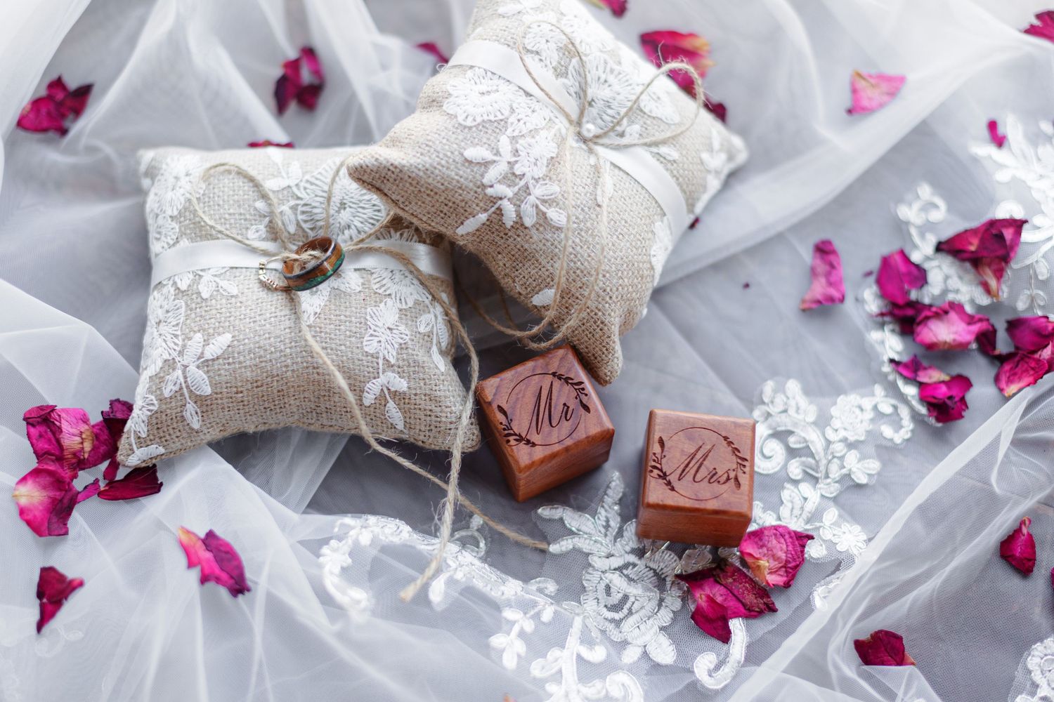 His and hers wooden ring boxes surrounded by ringbearer pillows, tulle, and rose petals.