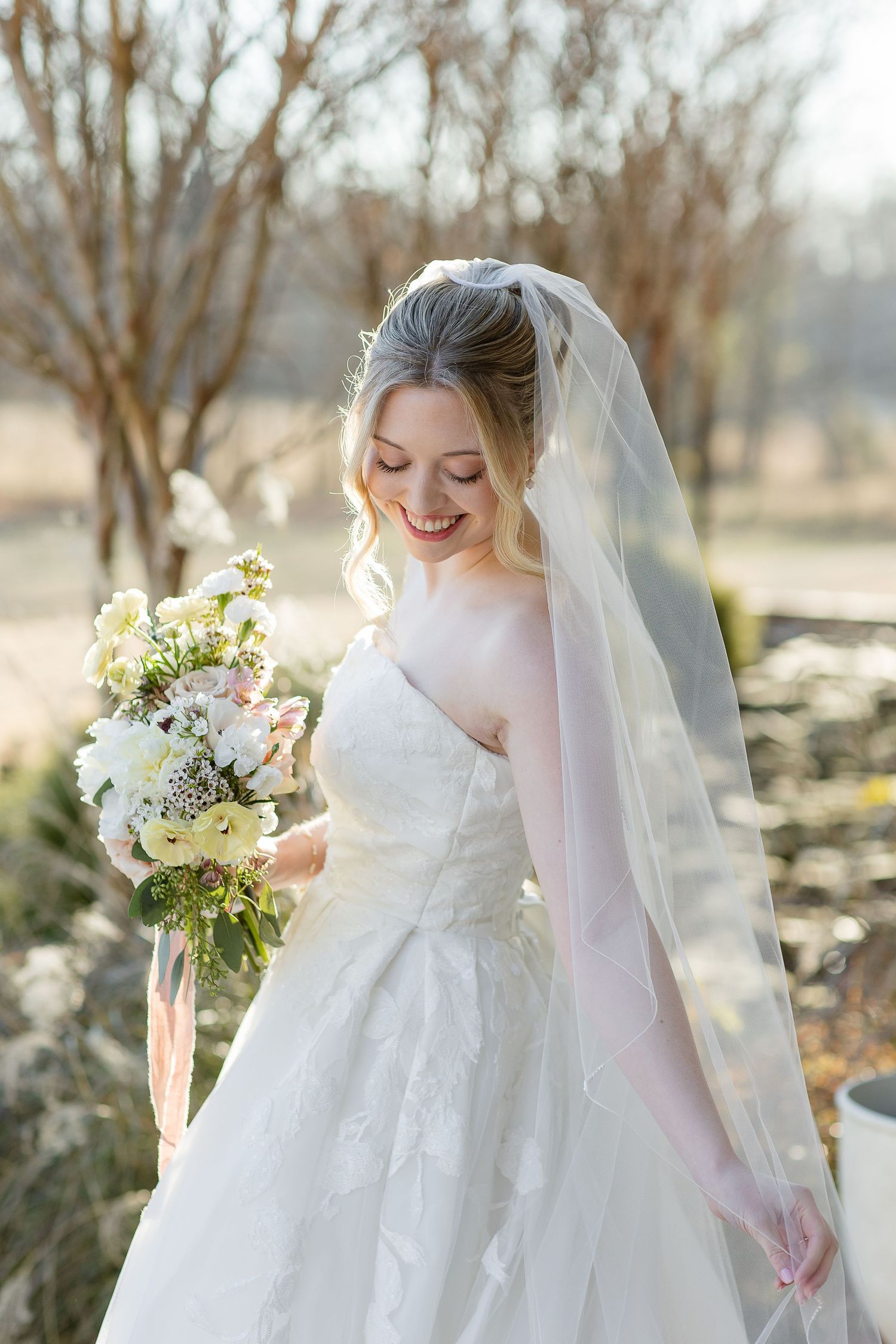 A beautiful bride flows her veil while smiling gently on a bright, sunny day. Image by Emily Joy, wedding photographer in Memphis.